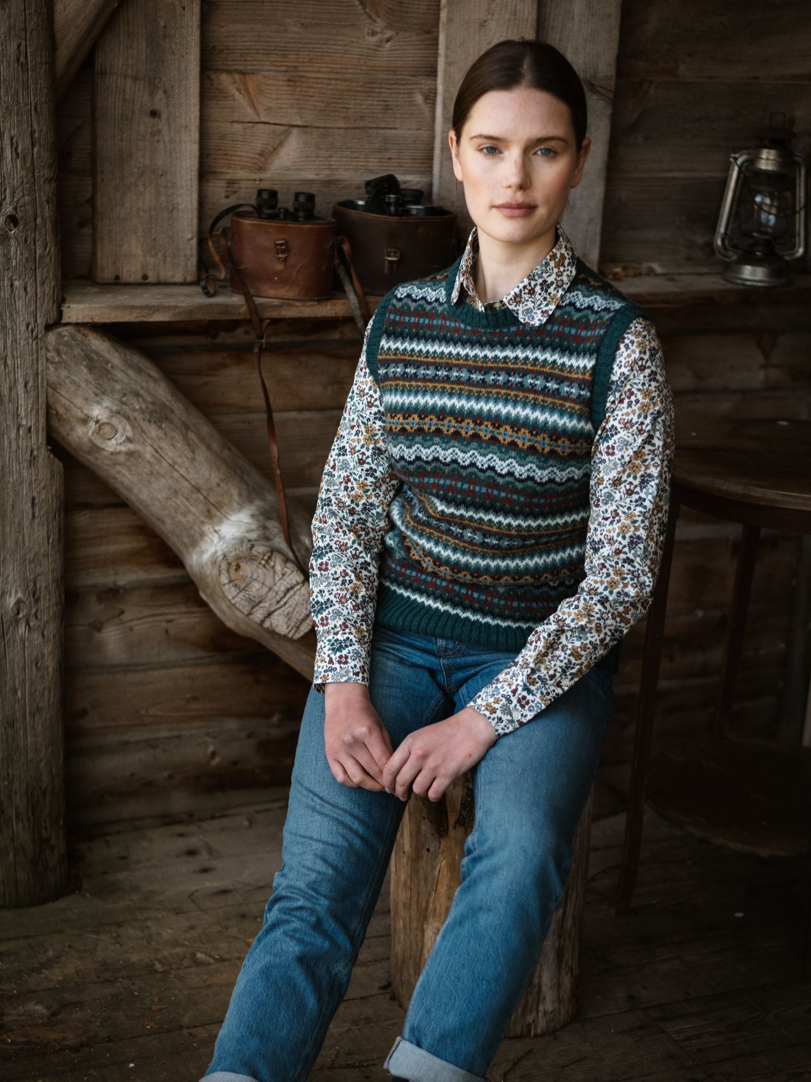 A person with brown hair in a low ponytail sits on a wooden stool in a rustic cabin, wearing a patterned sweater vest over the Campbell's of Beauly Liberty Print Cotton Shirt and blue jeans, with vintage lanterns and saddles behind them.