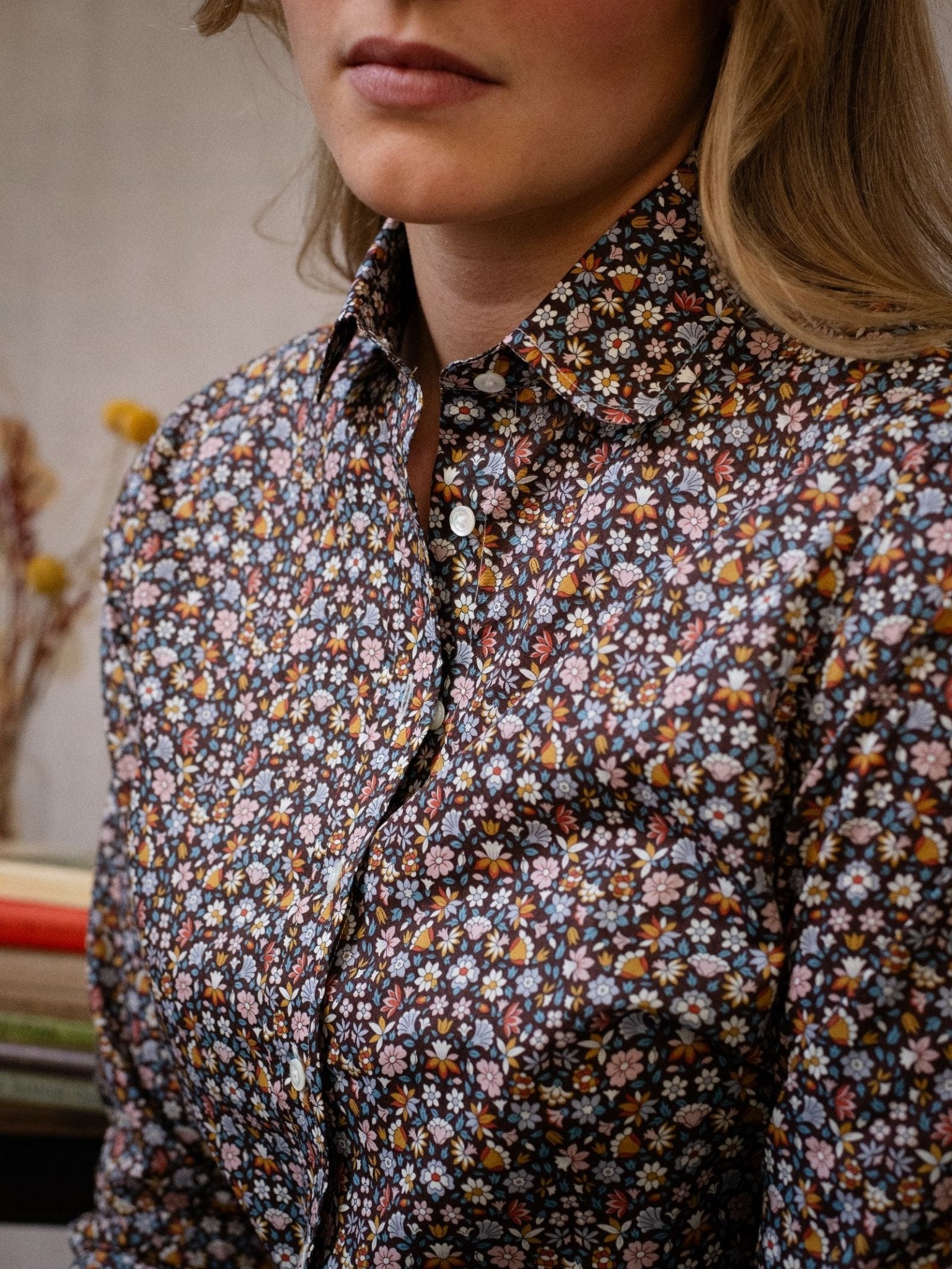 A person with light brown hair wears a Liberty Print Cotton Shirt by Campbell's of Beauly, featuring a small colorful floral pattern. Only their lower face and upper torso are visible against a blurred, muted background.