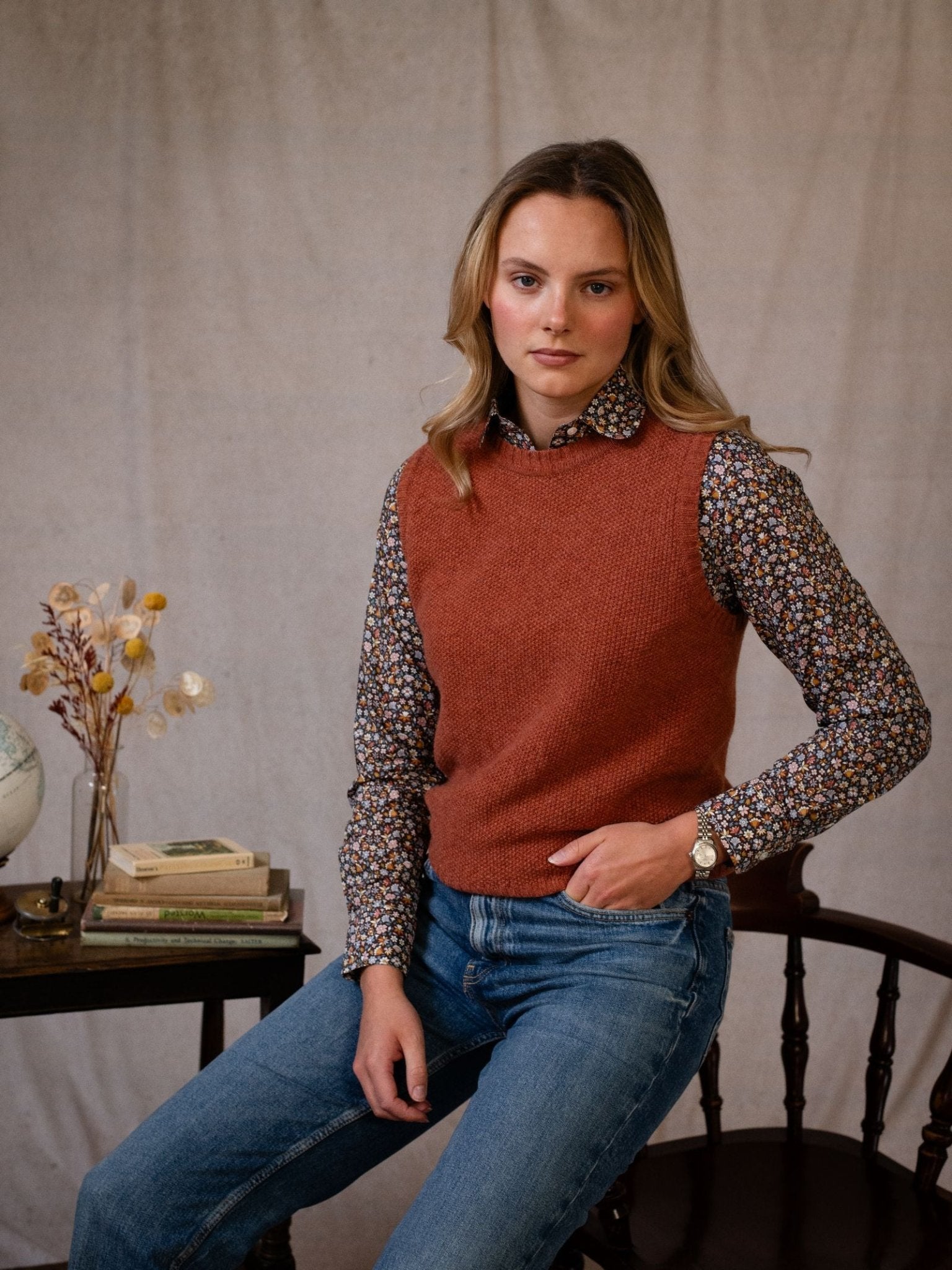 A woman with light brown hair wears a rust-colored knit vest over a Campbell's of Beauly Liberty Print Cotton Shirt and blue jeans, seated on a wooden chair by a table with books, a globe, and dried flowers against a neutral backdrop.