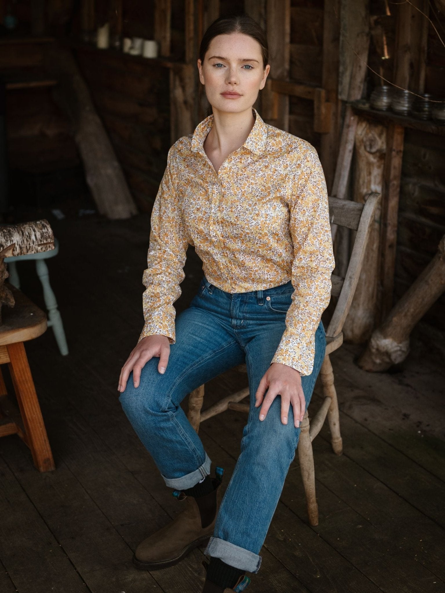 A woman with brown hair sits indoors on a wooden chair, wearing a yellow Liberty Print Cotton Shirt by Campbell's of Beauly, blue jeans, and brown boots. She faces the camera calmly in a rustic wood-paneled room.