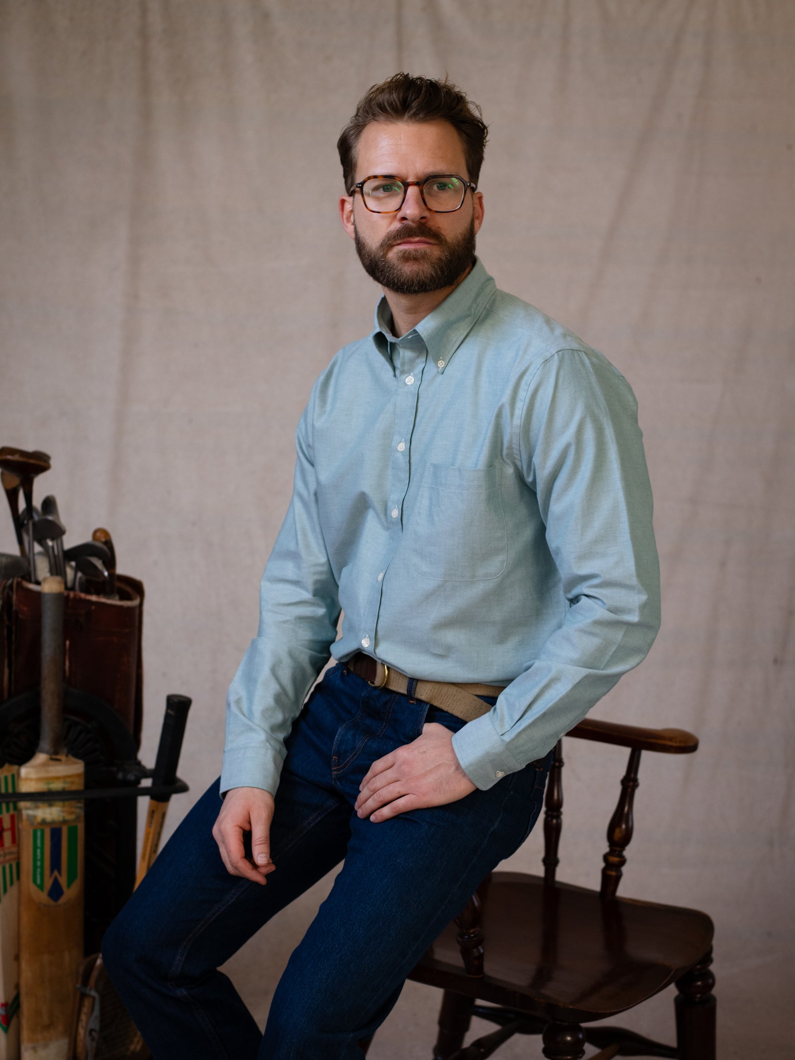 A bearded man in glasses sits on a wooden chair wearing the Campbell's of Beauly Oxford Button Down Shirt and jeans, with vintage golf clubs and a cricket bat beside him, all set against a neutral background.