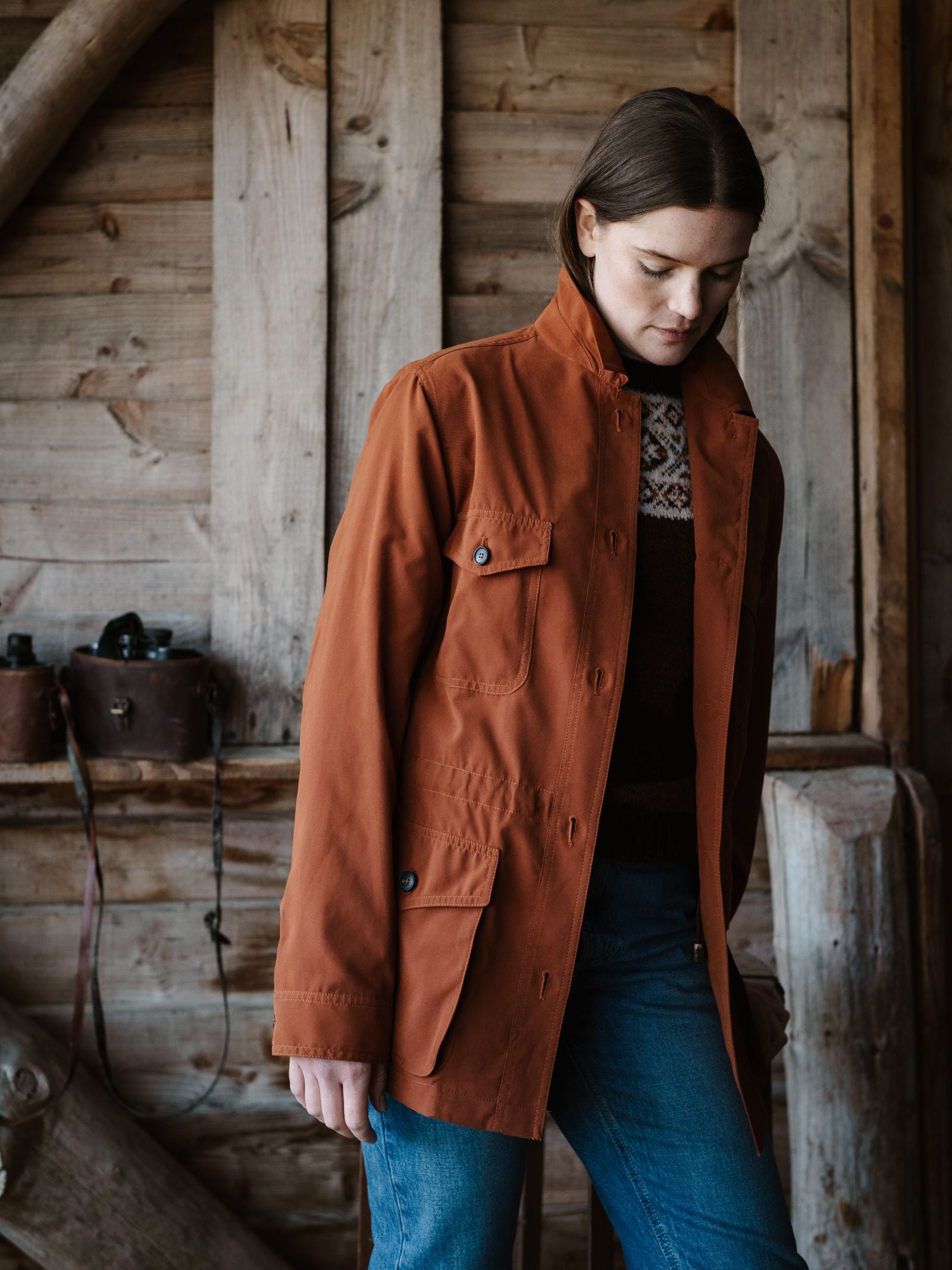 A woman with straight brown hair wears the Lightweight Aigas Field Jacket by Campbell's of Beauly over a patterned sweater and jeans, standing indoors against a rustic wooden wall with a vintage camera on a shelf behind her.