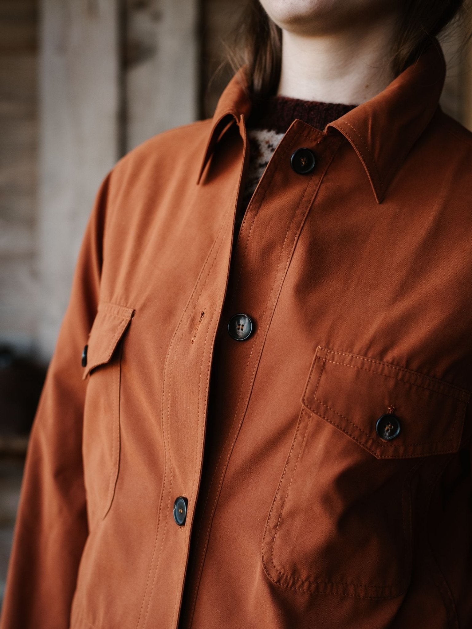 A person wearing Campbell's of Beauly's Lightweight Aigas Field Jacket in rust, featuring two chest pockets and crafted from Belseta fabric, stands indoors against a blurred wooden background with only the lower part of their face visible.