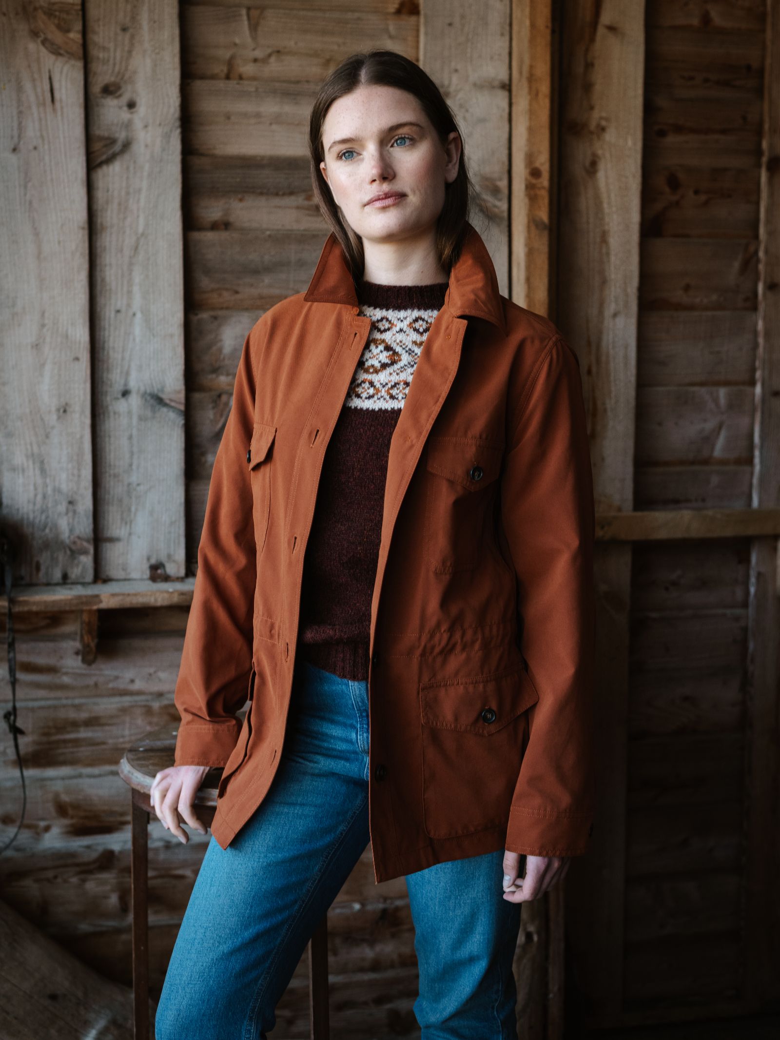 A woman stands indoors against a rustic wooden wall, wearing the Lightweight Aigas Field Jacket in rust by Campbell's of Beauly over a patterned sweater and blue jeans, looking slightly to the side with a calm expression.