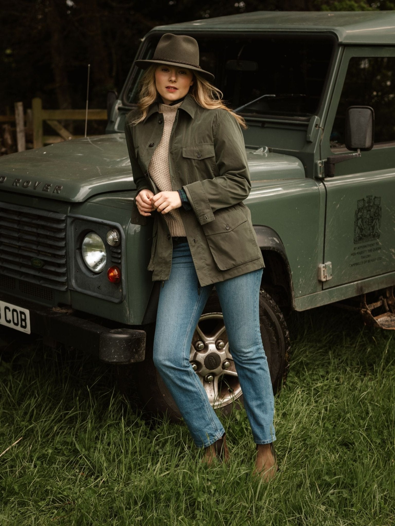A woman in a Campbell's of Beauly Lightweight Aigas Field Jacket, hat, and blue jeans leans against a green Land Rover Defender on grass, with trees and a wooden fence behind her.