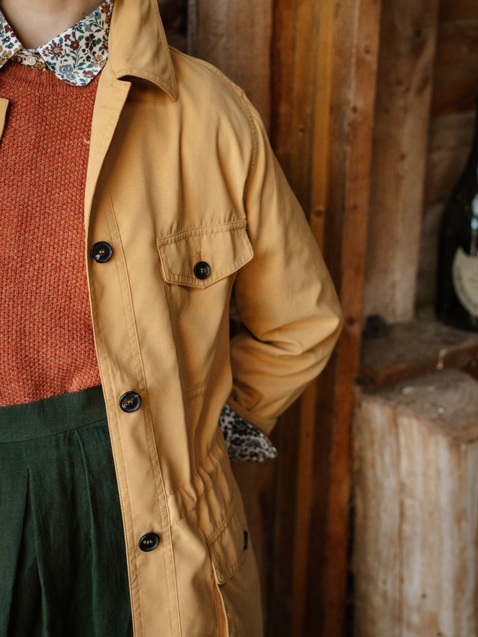 A person in a Campbell's of Beauly Lightweight Aigas Field Jacket in mustard yellow, layered over a rust sweater, floral collared shirt, and high-waisted green pants, stands indoors by wooden walls with their face hidden.