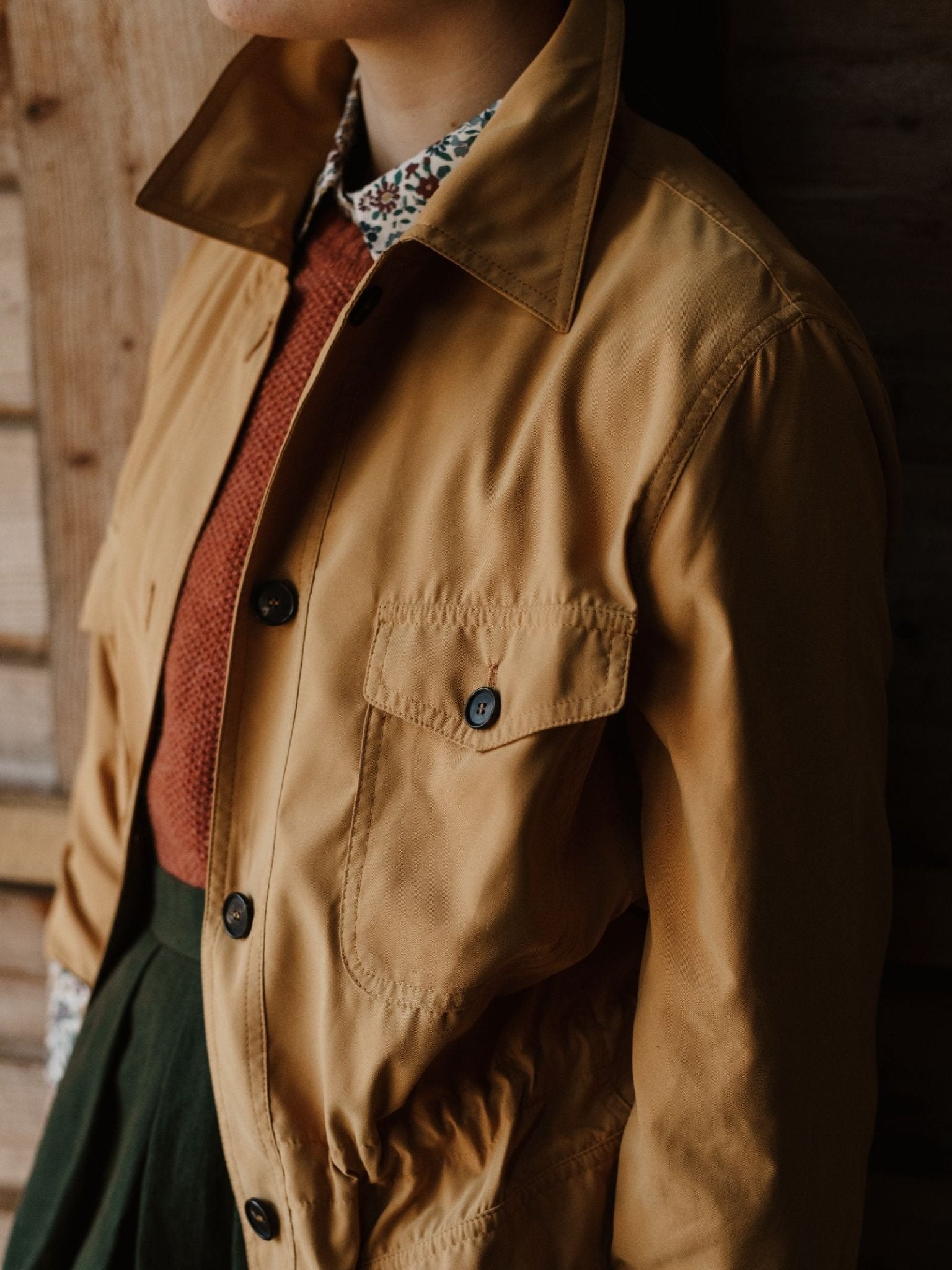 A person wears the Campbell's of Beauly Lightweight Aigas Field Jacket in tan over a rust sweater and a floral-collared shirt, standing against a wooden wall. The outfit is shown partially from neck to waist.