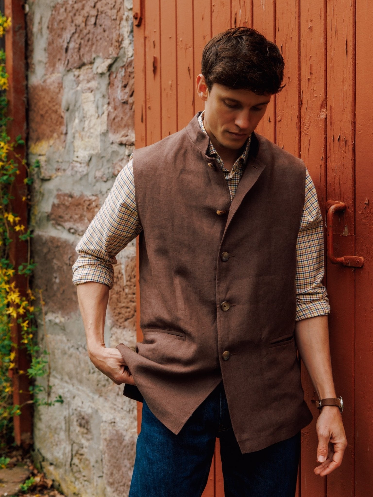 A man stands by a red wooden door and stone wall, wearing Campbell's of Beauly Linen Nehru Gilet over a checked shirt with rolled sleeves and blue jeans—a stylish summer layer. He looks down, holding the edge of his vest.