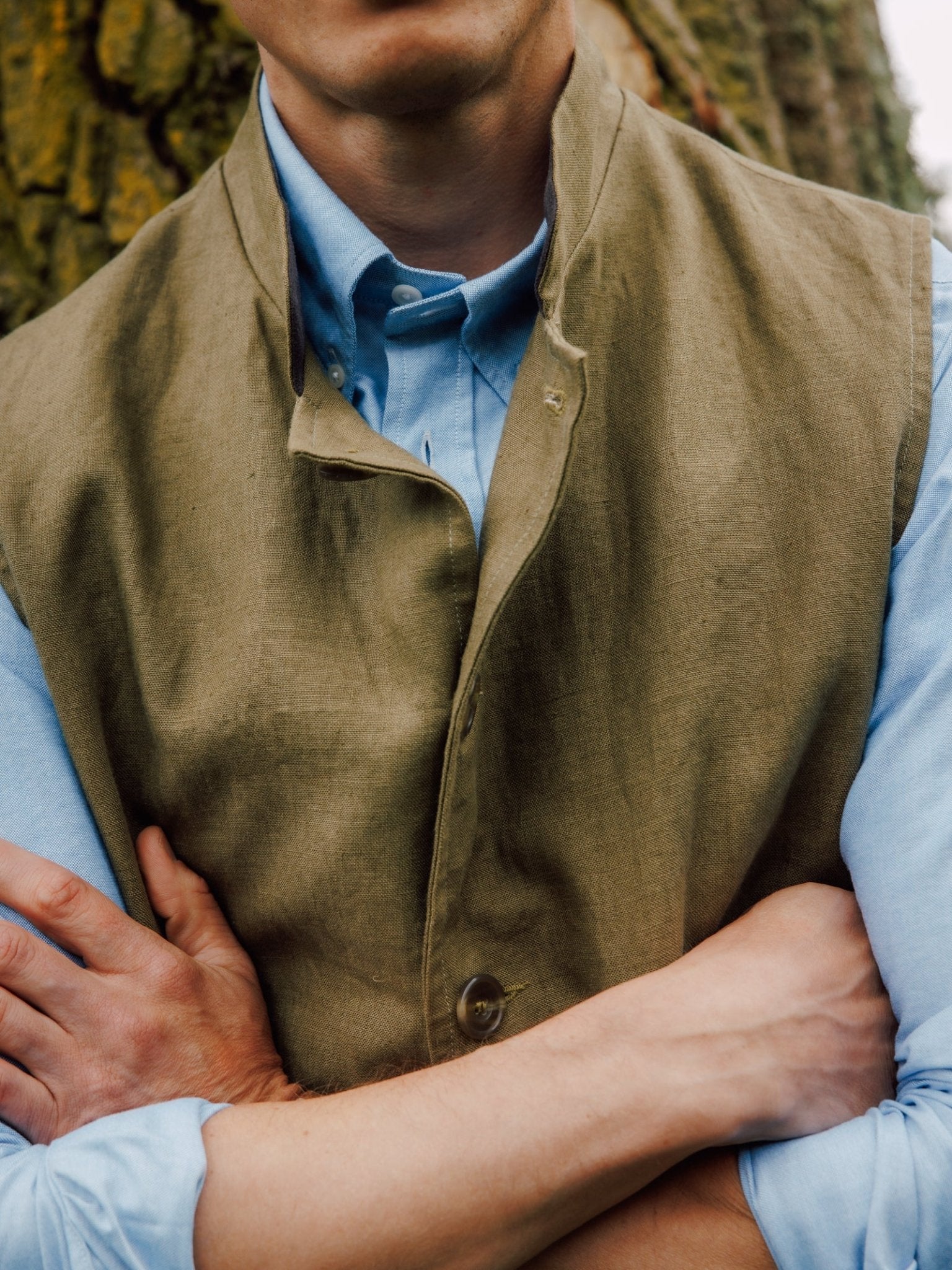 A person stands with arms crossed, wearing the Campbell's of Beauly Linen Nehru Gilet over a light blue shirt. A tree trunk with textured bark appears in the background.