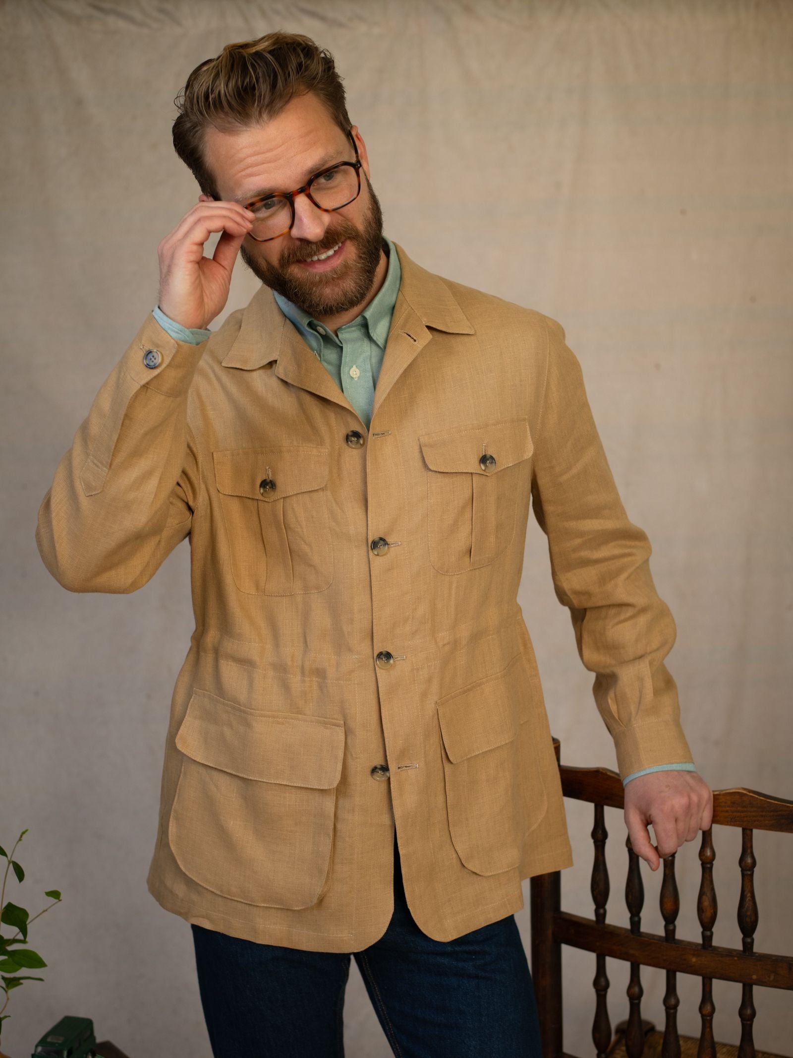 A man with glasses and a beard wears a tan Linen Safari Jacket by Campbell's of Beauly over a green shirt, standing indoors and adjusting his glasses with a slight smile. A wooden chair and plant appear in the background.