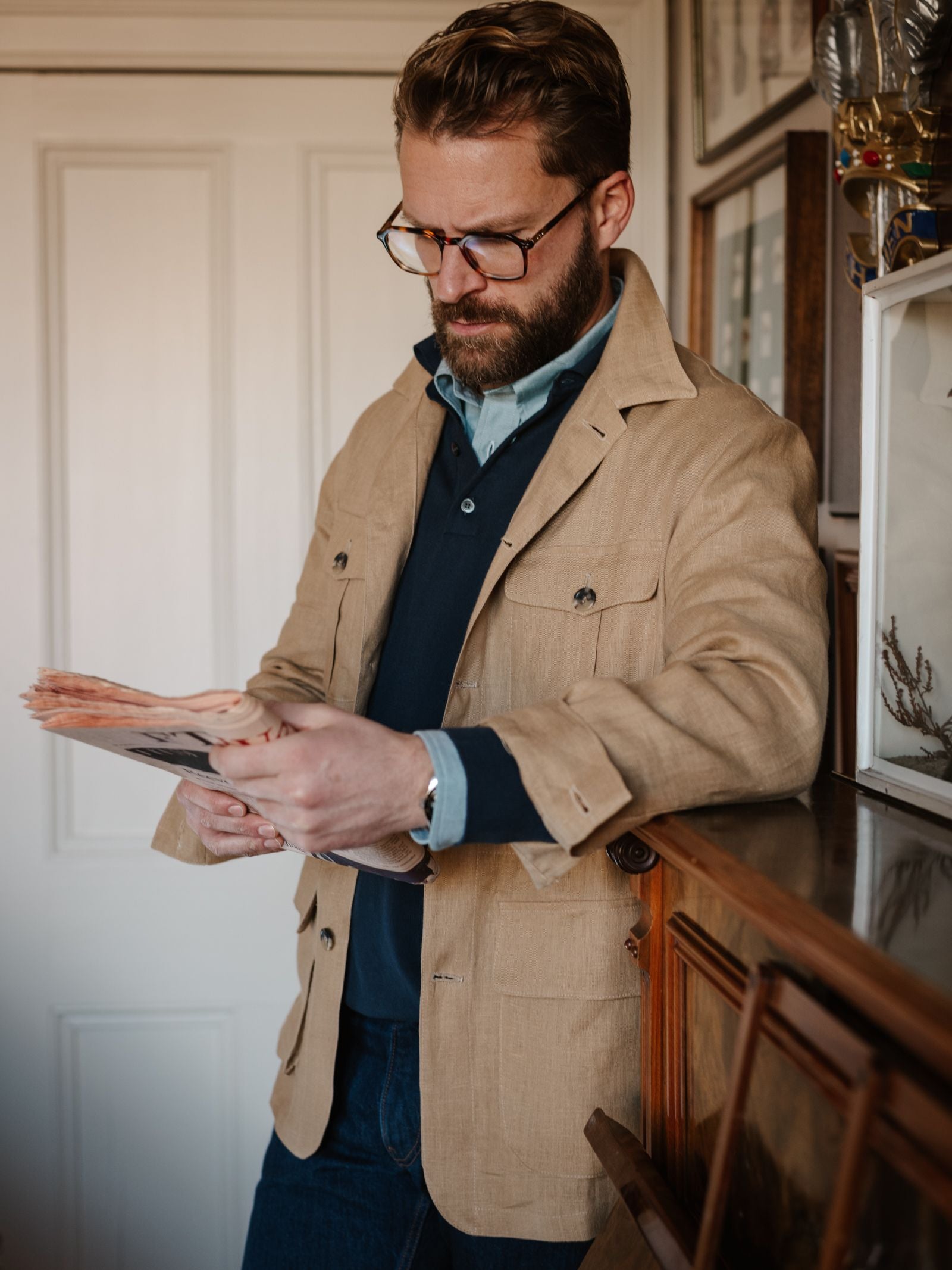 A bearded man in glasses wears the Campbell's of Beauly Linen Safari Jacket in light brown over a blue shirt and dark sweater, standing indoors by a wooden cabinet as he reads a newspaper.