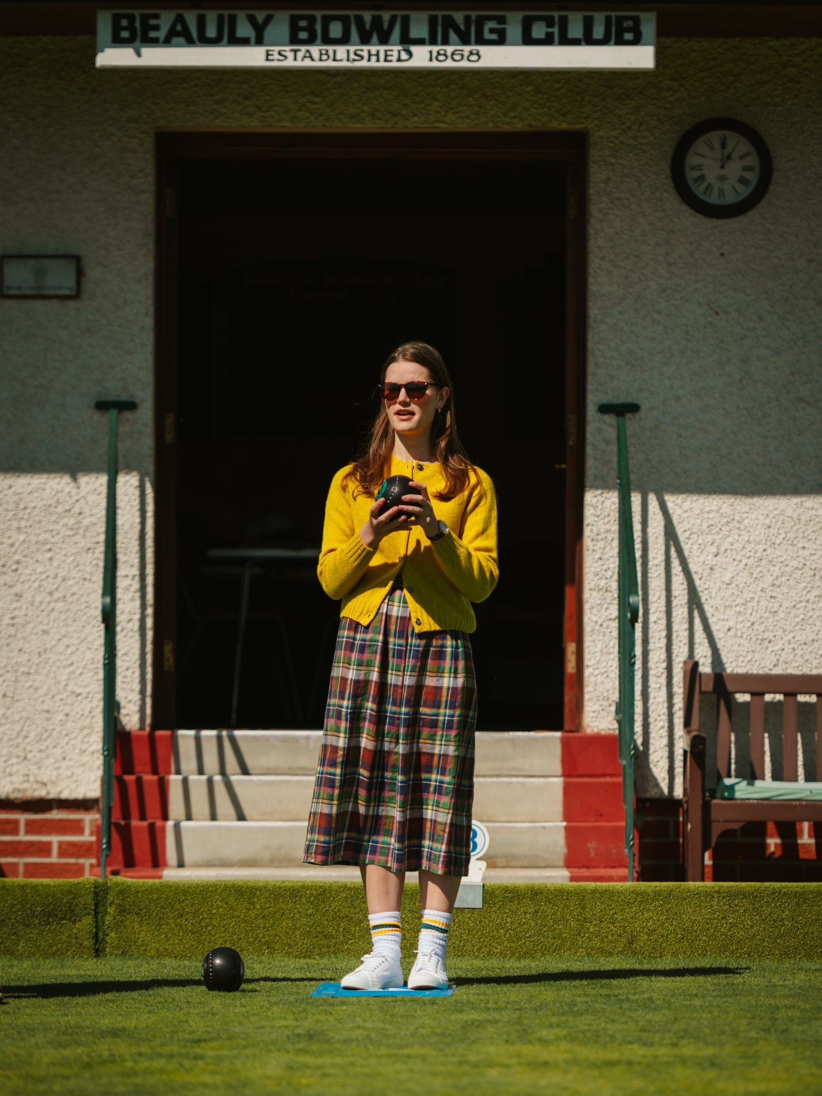 A woman wearing a Campbell's of Beauly Linen Skirt, yellow cardigan, and sunglasses stands on a bowling green holding a trophy, with the "Beauly Bowling Club" sign visible above the doorway behind her.
