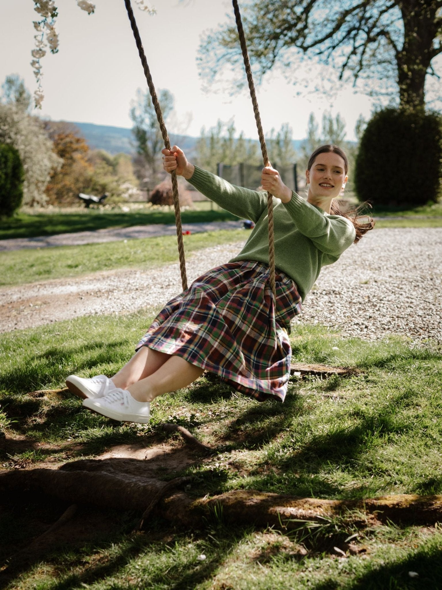 A young woman smiles as she swings outdoors, wearing a Campbell's of Beauly Linen Skirt, paired with a green sweater and white sneakers amid grass, trees, and sunlight.
