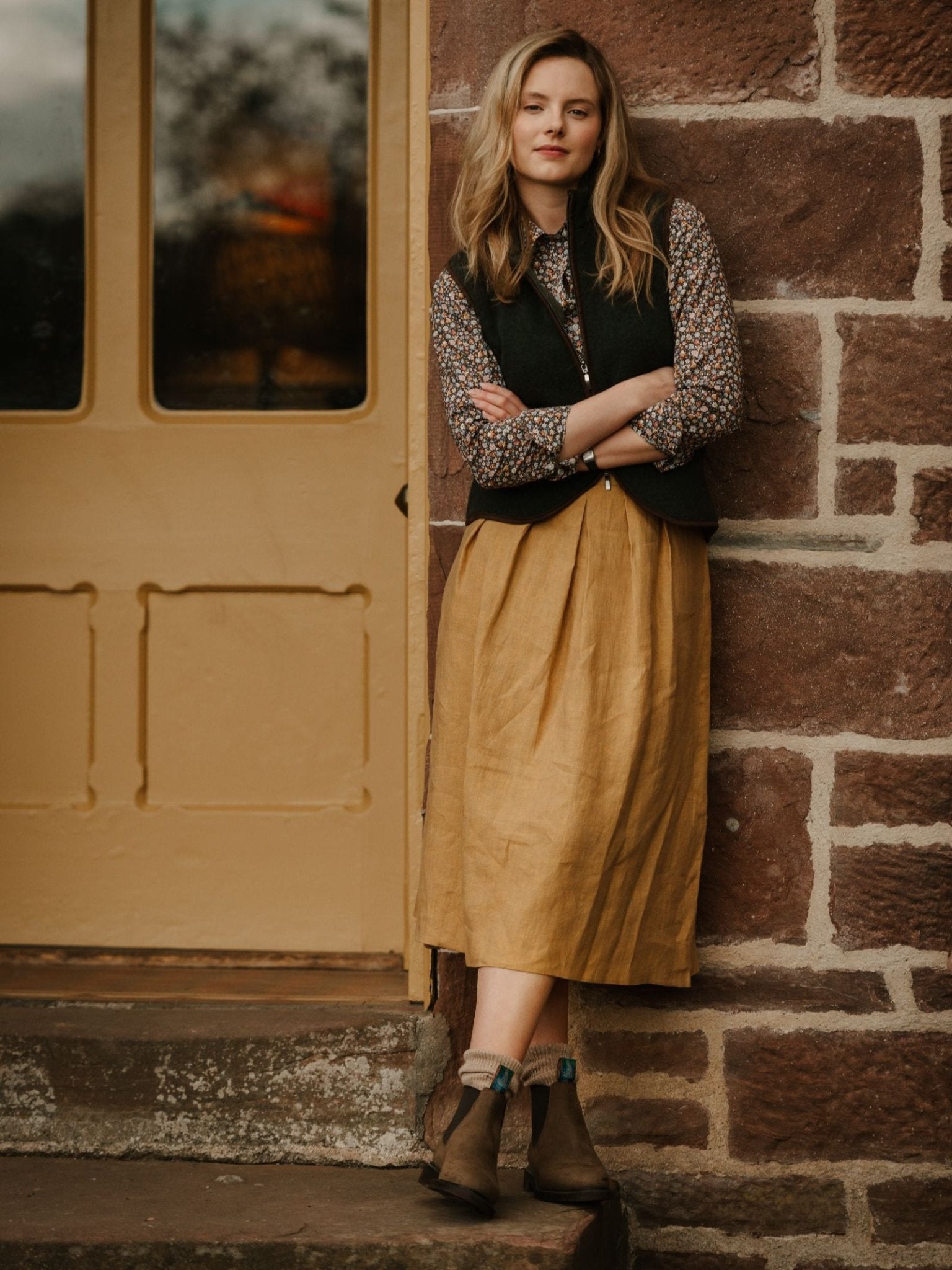 A woman with blonde hair leans against a brick wall on a stone doorstep, wearing the Campbell's of Beauly Linen Skirt, a floral blouse, dark vest, and brown ankle boots. Behind her is a yellow door.
