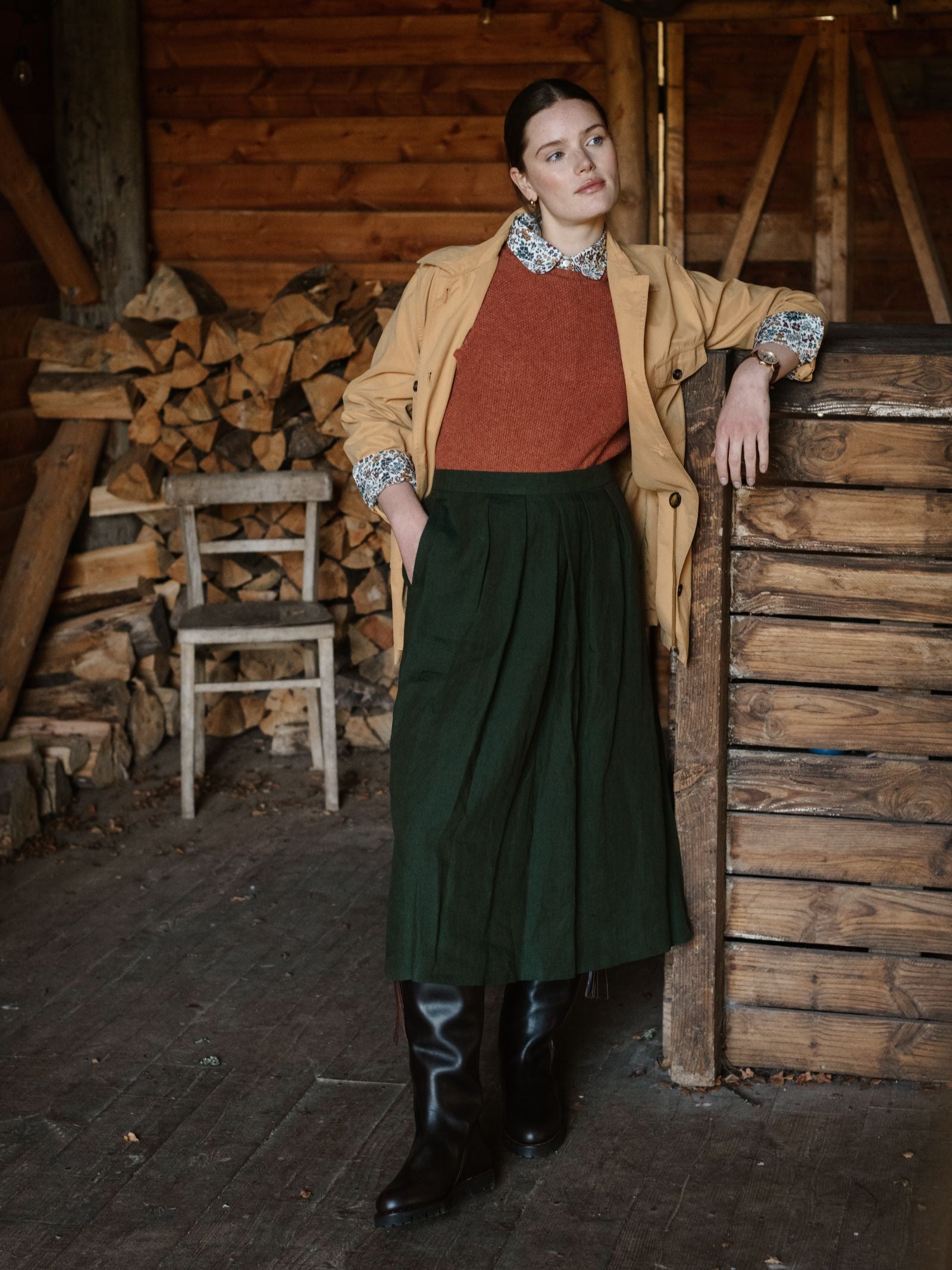 A woman in a mustard jacket, rust sweater, and Campbell's of Beauly Linen Skirt with black boots leans on a wooden wall in a rustic cabin, with stacked firewood and a wooden chair behind her.