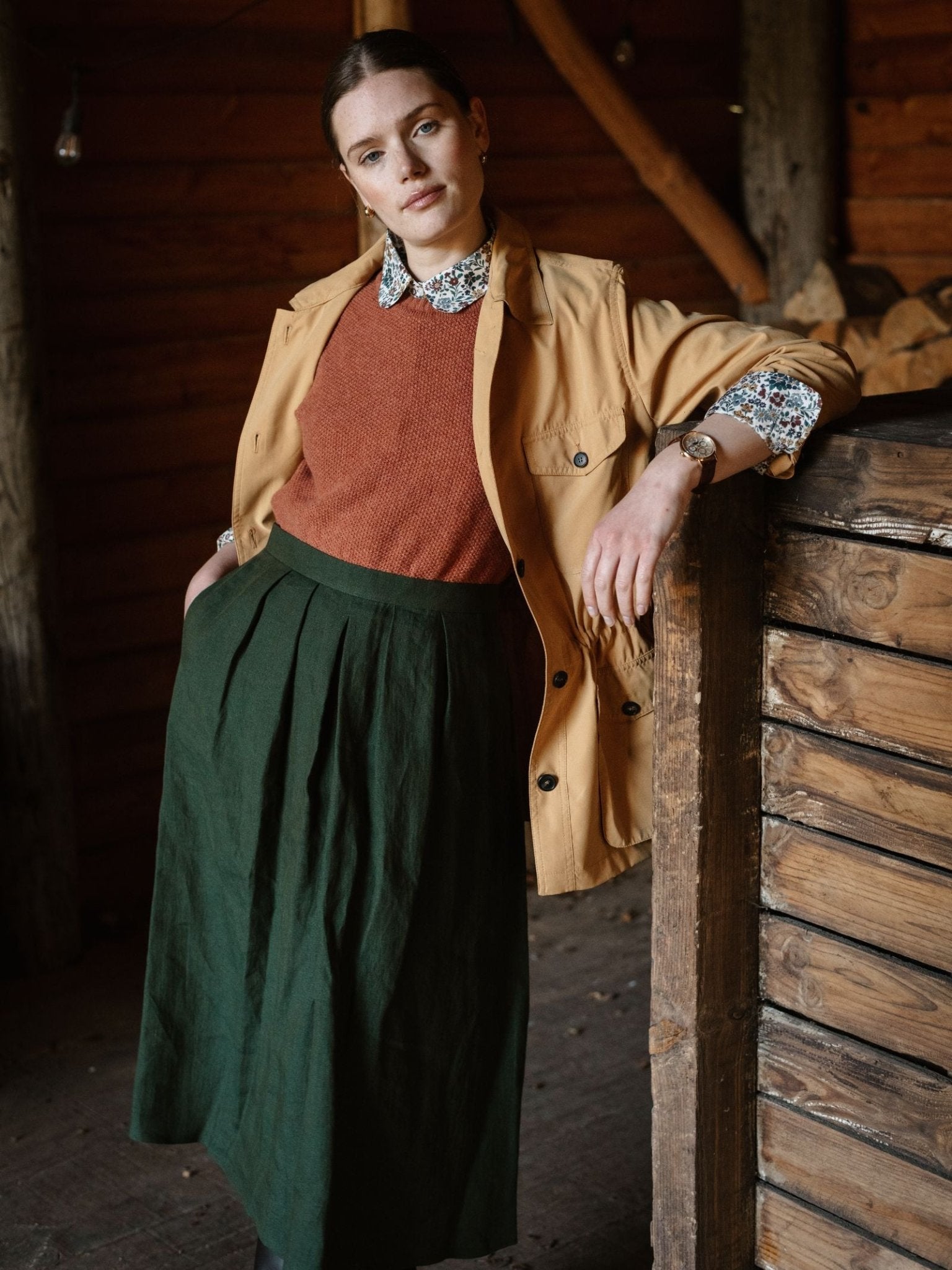 A woman stands indoors by a wooden railing, wearing Campbell's of Beauly Linen Skirt in deep green, paired with a tan jacket, rust sweater, and floral collared shirt. Wooden walls and stacked logs form the background.