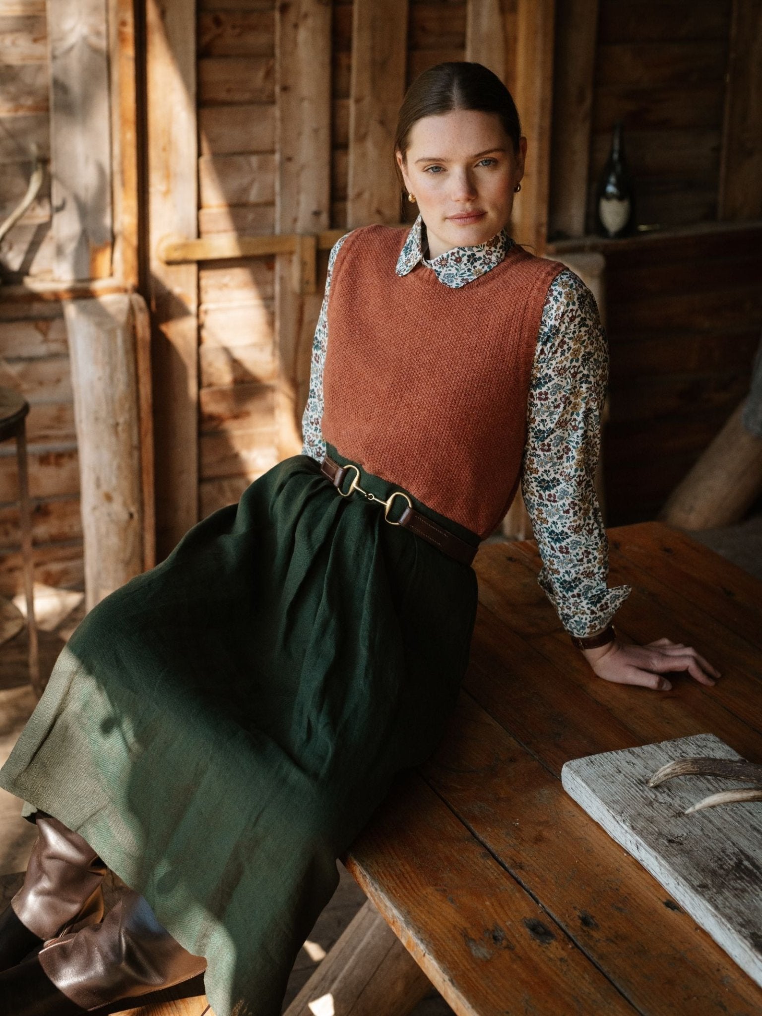 A woman wearing the Campbell's of Beauly Linen Skirt sits on a rustic wooden table in a cozy, sunlit cabin, looking calmly at the camera.