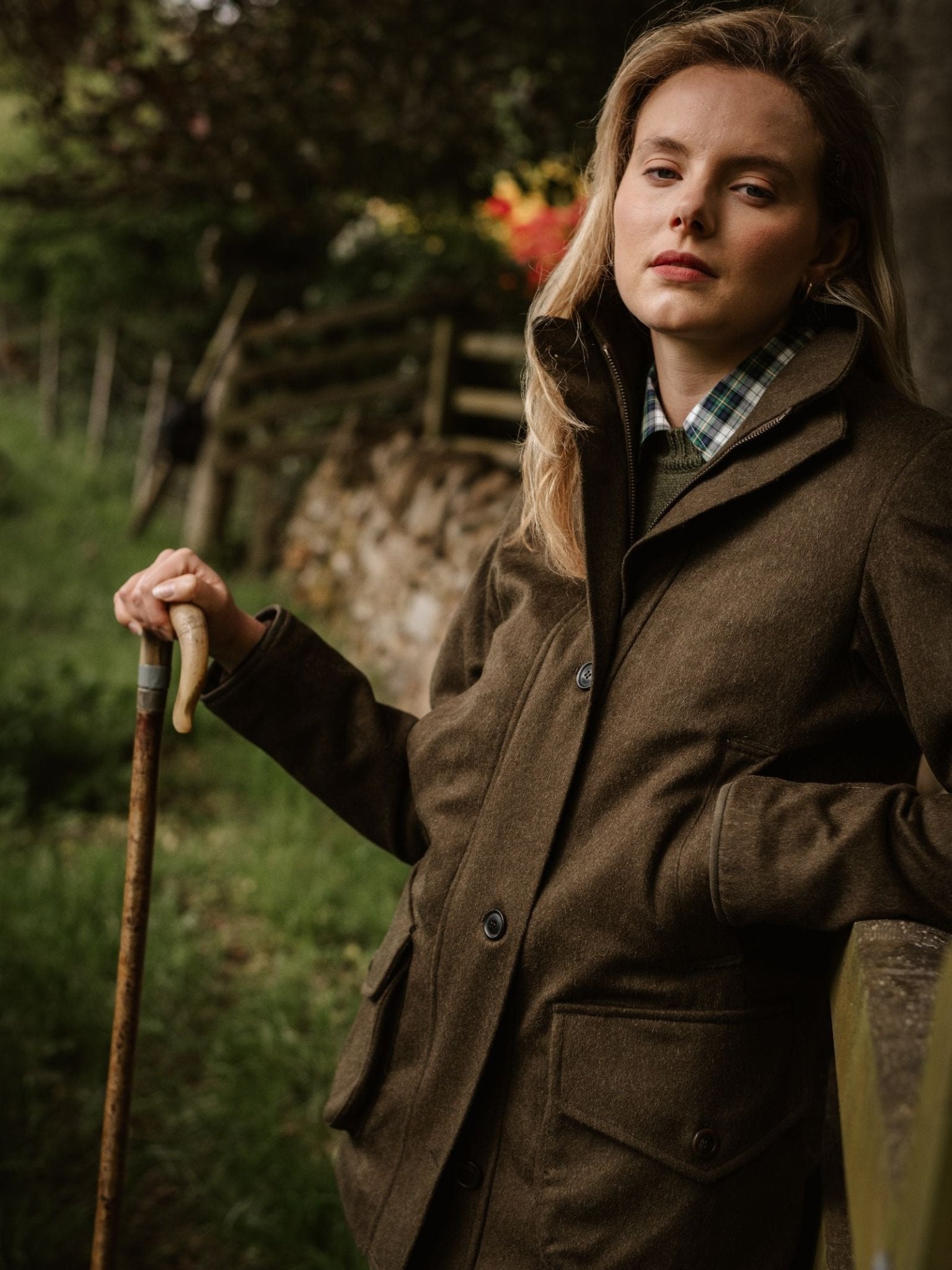 Wearing the Campbell's of Beauly Loden Green Field Coat, a woman stands by a fence with greenery and a stone wall behind her, holding a walking stick and gazing calmly at the camera.