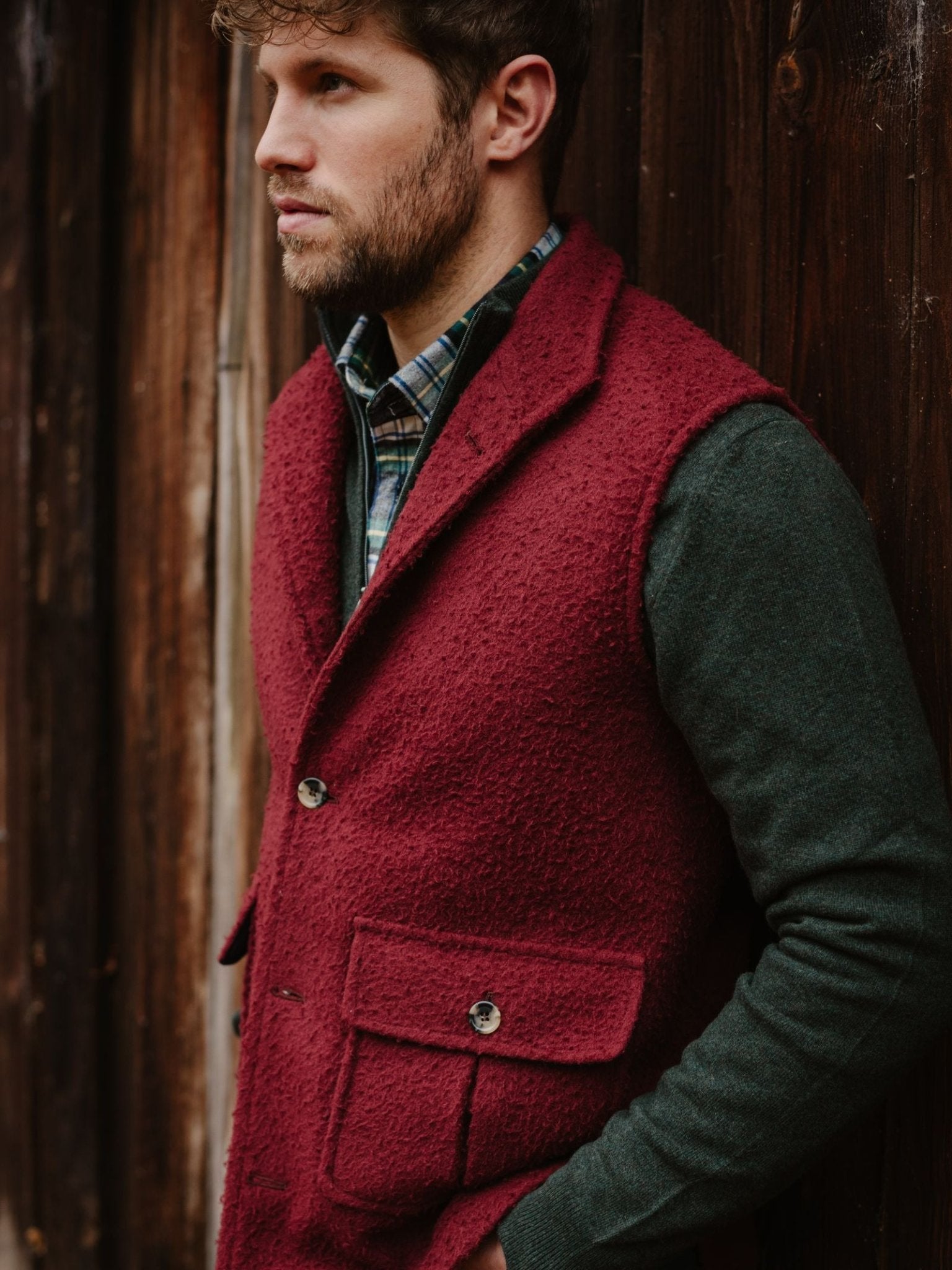 A bearded man in a green plaid shirt and a red Campbell’s of Beauly Casentino Fleece Gilet stands by a wooden wall, gazing thoughtfully to the side.