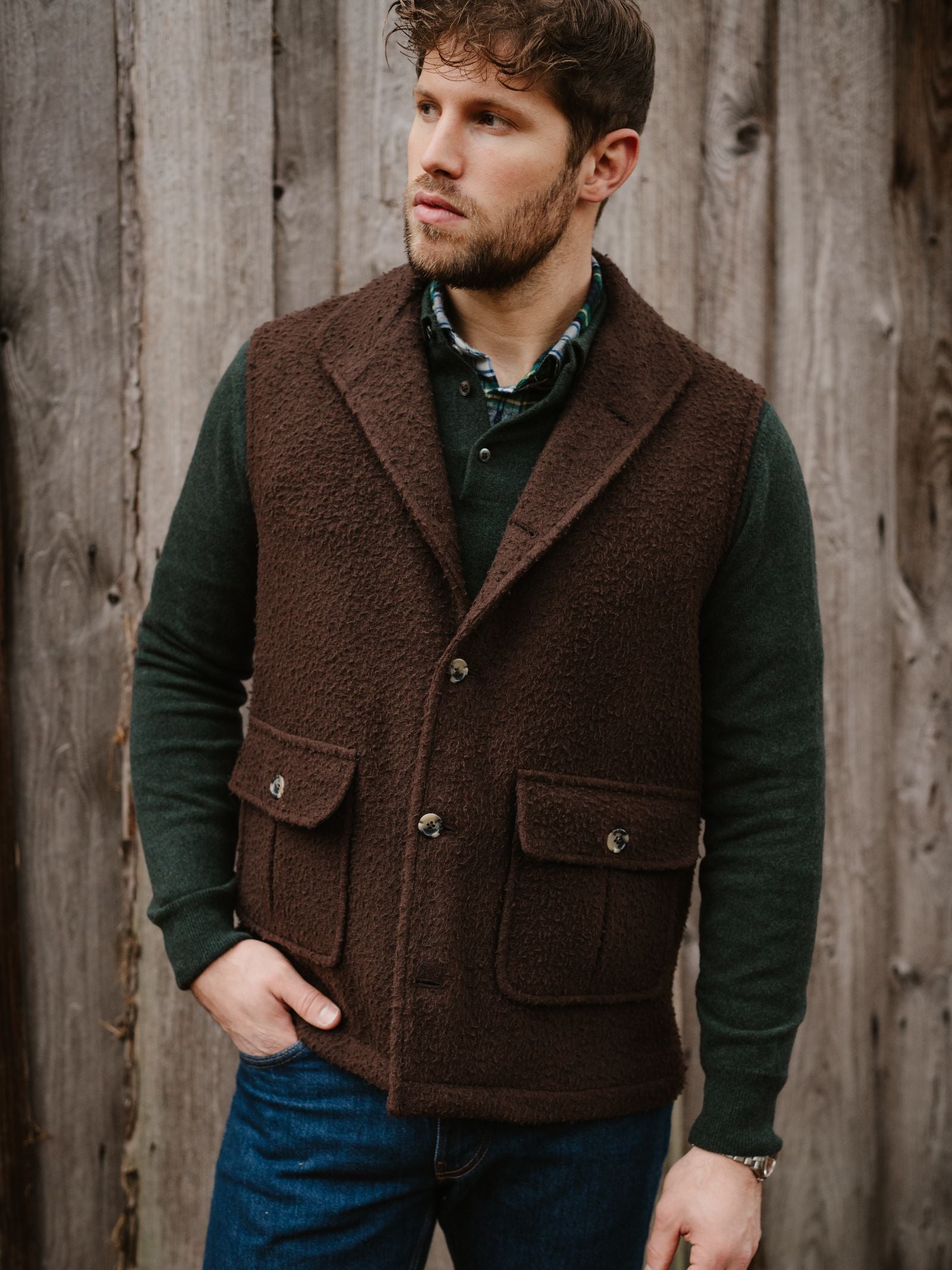A man with short brown hair and a trimmed beard stands by a wooden wall, wearing blue jeans, a dark green shirt, and the Campbell's of Beauly Casentino Fleece Gilet with pockets. He looks to the side with his hand in his pocket.