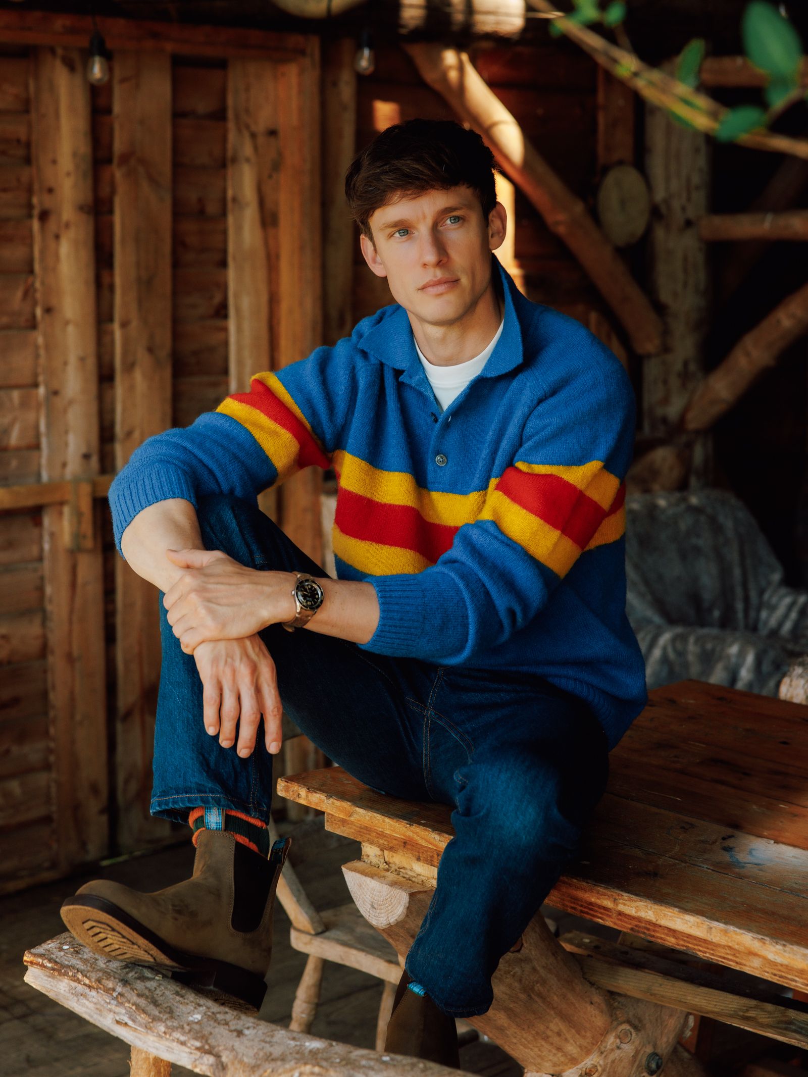A man in a blue sweater, jeans, and the Men's R.M.Williams Highlander Boot from the R.M.Williams x Campbell's of Beauly collaboration sits on a rustic bench in a shed, resting one arm on his knee and looking at the camera with a relaxed expression.