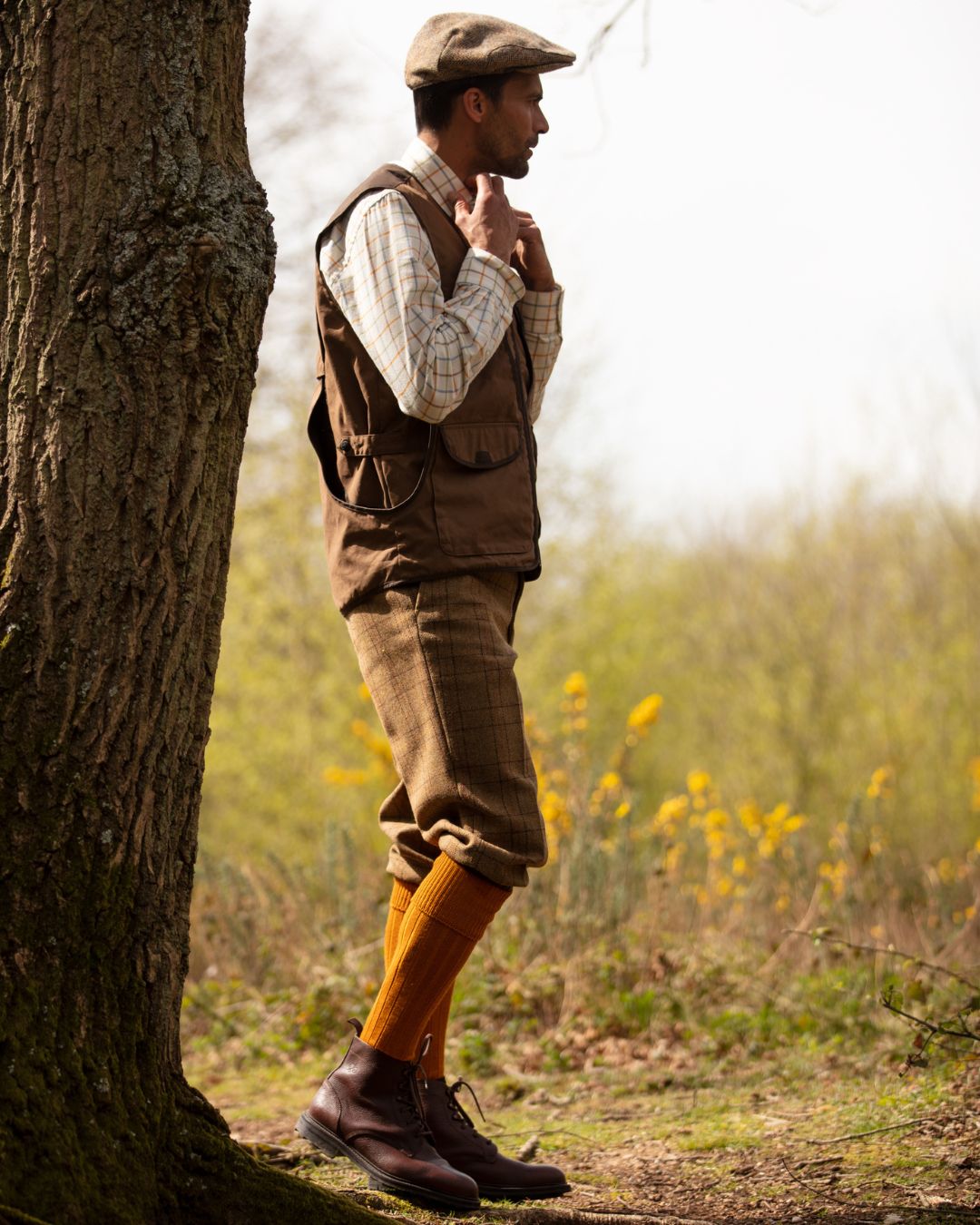 A man in vintage outdoor attire stands by a tree, wearing Campbell's of Beauly Scarba Shooting Socks in orange with brown boots, a flat cap, checked shirt, brown vest, and plaid knickerbocker trousers. Wildflowers and trees fill the background.