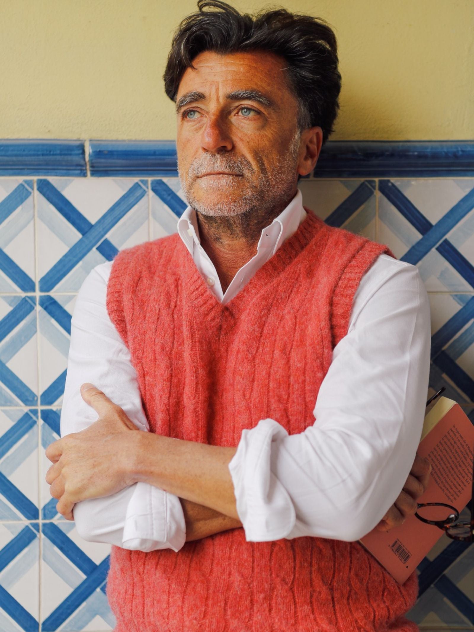 A man with gray hair and a beard, wearing a white shirt and the Shetland Cable Slipover by Campbell's of Beauly, stands against a blue-and-white tiled wall with arms crossed, holding a book, looking thoughtful.