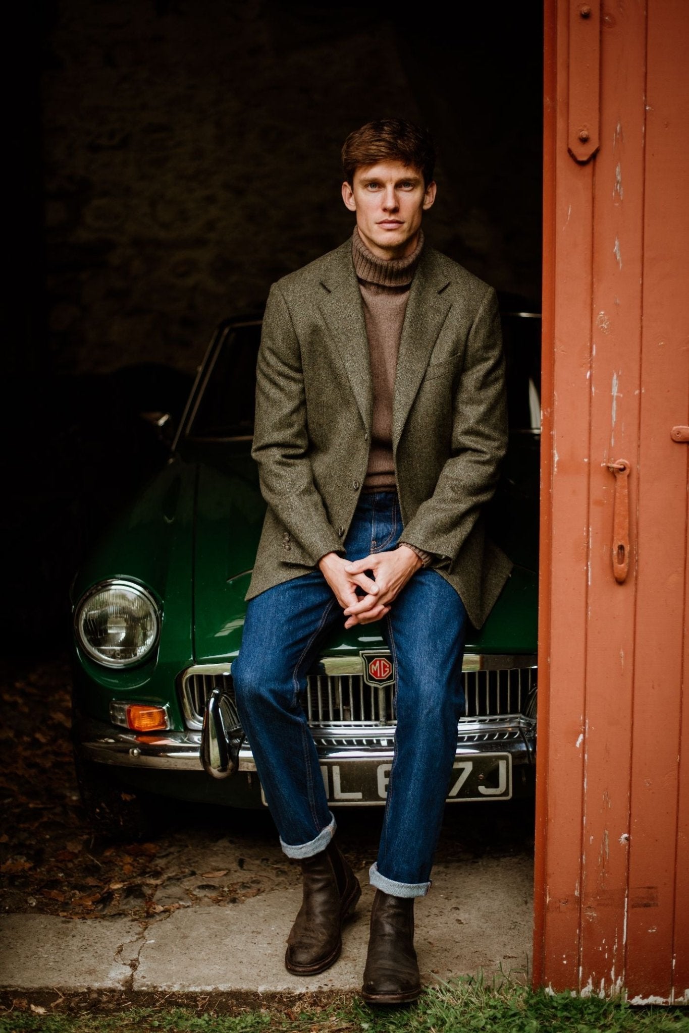A man wearing the Campbell's of Beauly Auld Stock House Jacket sits on the hood of a classic green MG, partially inside a garage with an open red door.