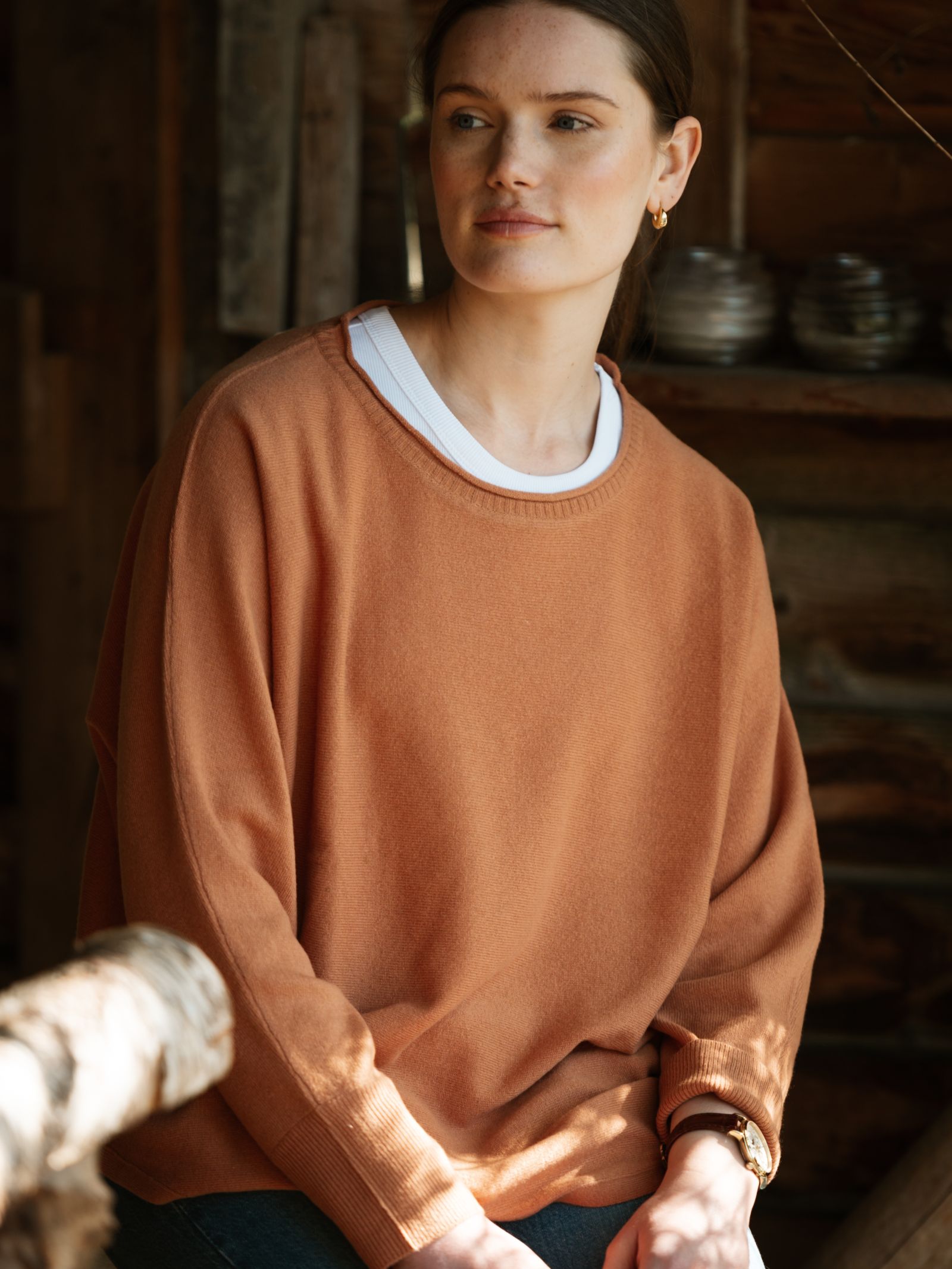 A woman with brown hair in a ponytail sits indoors by rustic wooden walls, softly lit by natural light, thoughtfully gazing aside. She wears the Campbell's of Beauly Merino Cashmere Boat Neck Jumper over a white shirt.