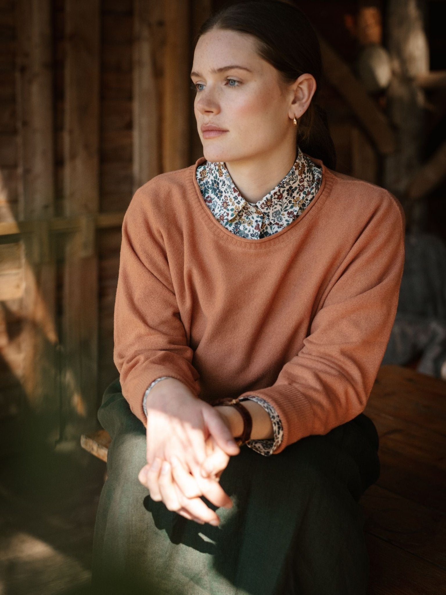 A woman with brown hair sits indoors, wearing the Merino Cashmere Boat Neck Jumper by Campbell's of Beauly over a floral collared shirt. Warm light highlights her thoughtful expression as she rests her hands in her lap.