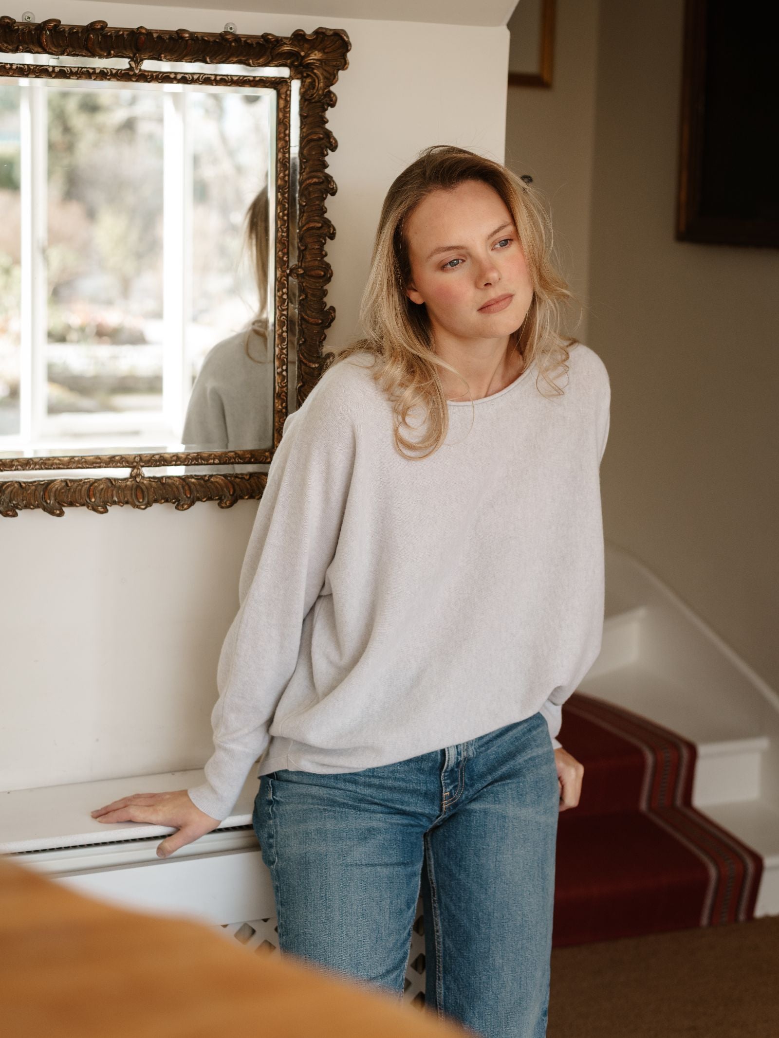 A blonde woman in blue jeans wears the Merino Cashmere Boat Neck Jumper by Campbell's of Beauly, leaning against a white wall by the stairs. A decorative mirror on the wall beside her reflects part of the room.