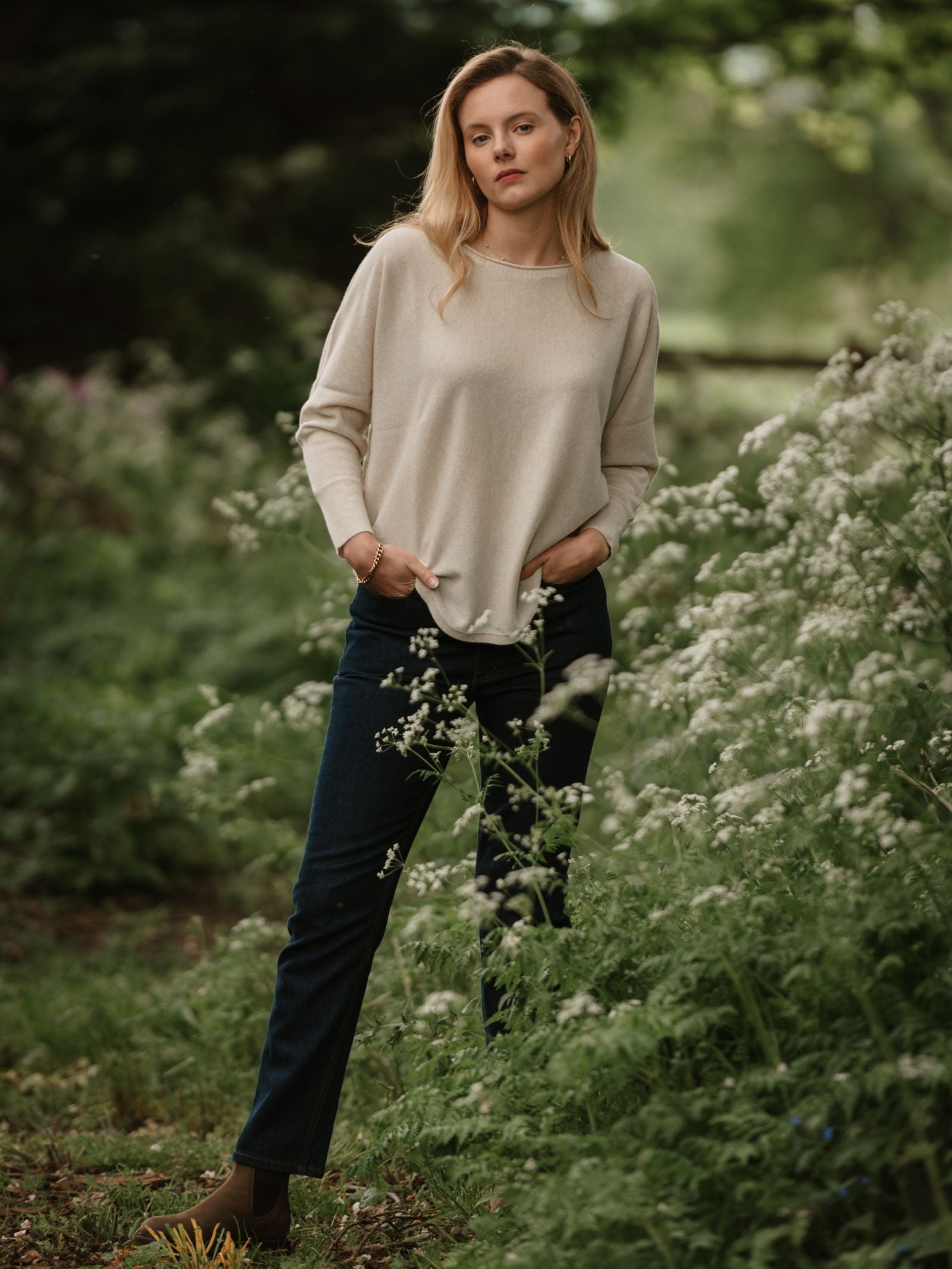 In a lush green outdoor setting, a person wears the Campbell’s of Beauly Merino Cashmere Boat Neck Jumper in beige, paired with dark jeans and brown boots, hands in pockets and a calm expression amid tall plants and white wildflowers.