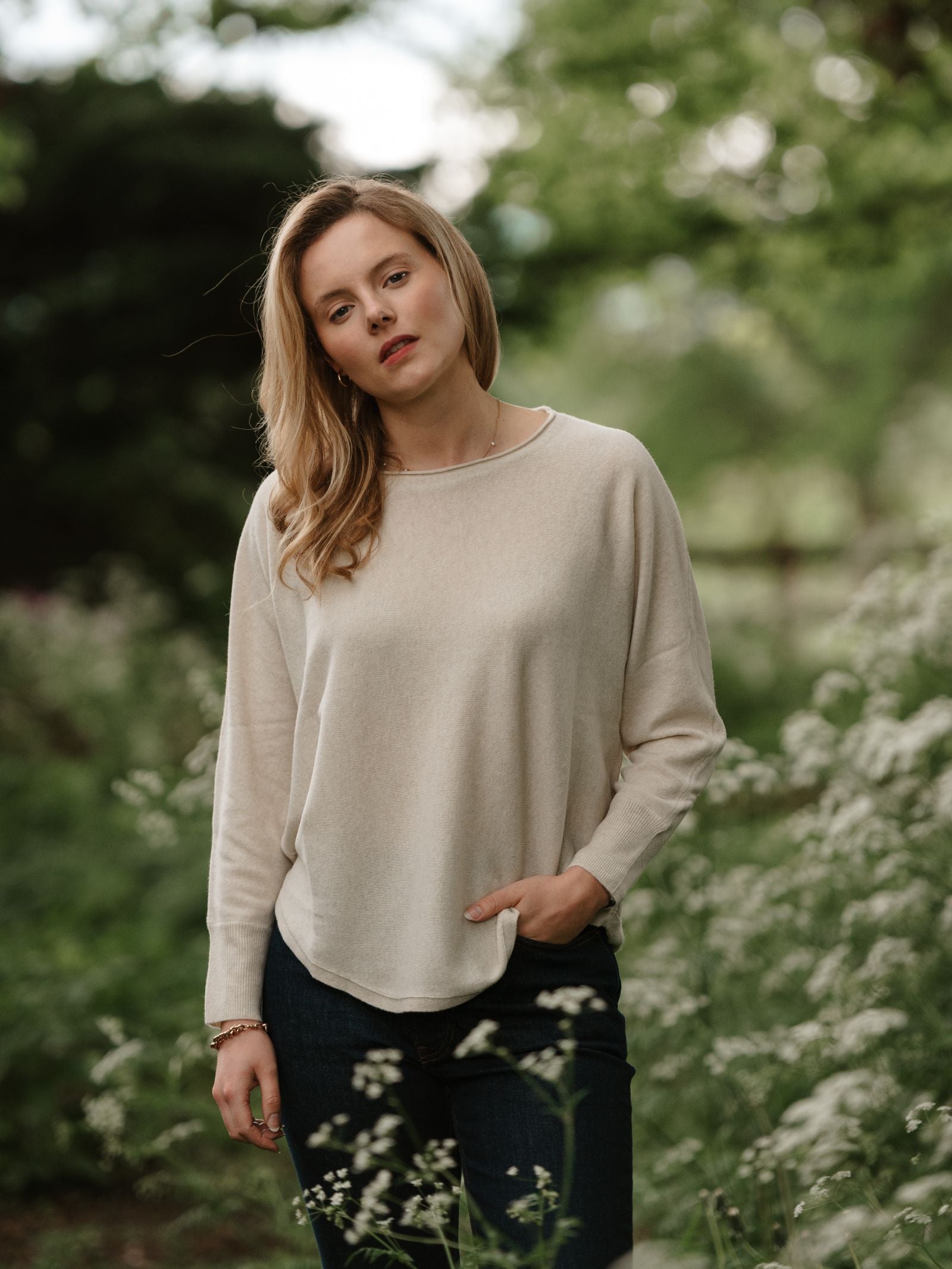 A blonde woman wears the Campbell's of Beauly Merino Cashmere Boat Neck Jumper in cream with dark jeans, standing outdoors among lush greenery and wildflowers, looking at the camera with a neutral expression.