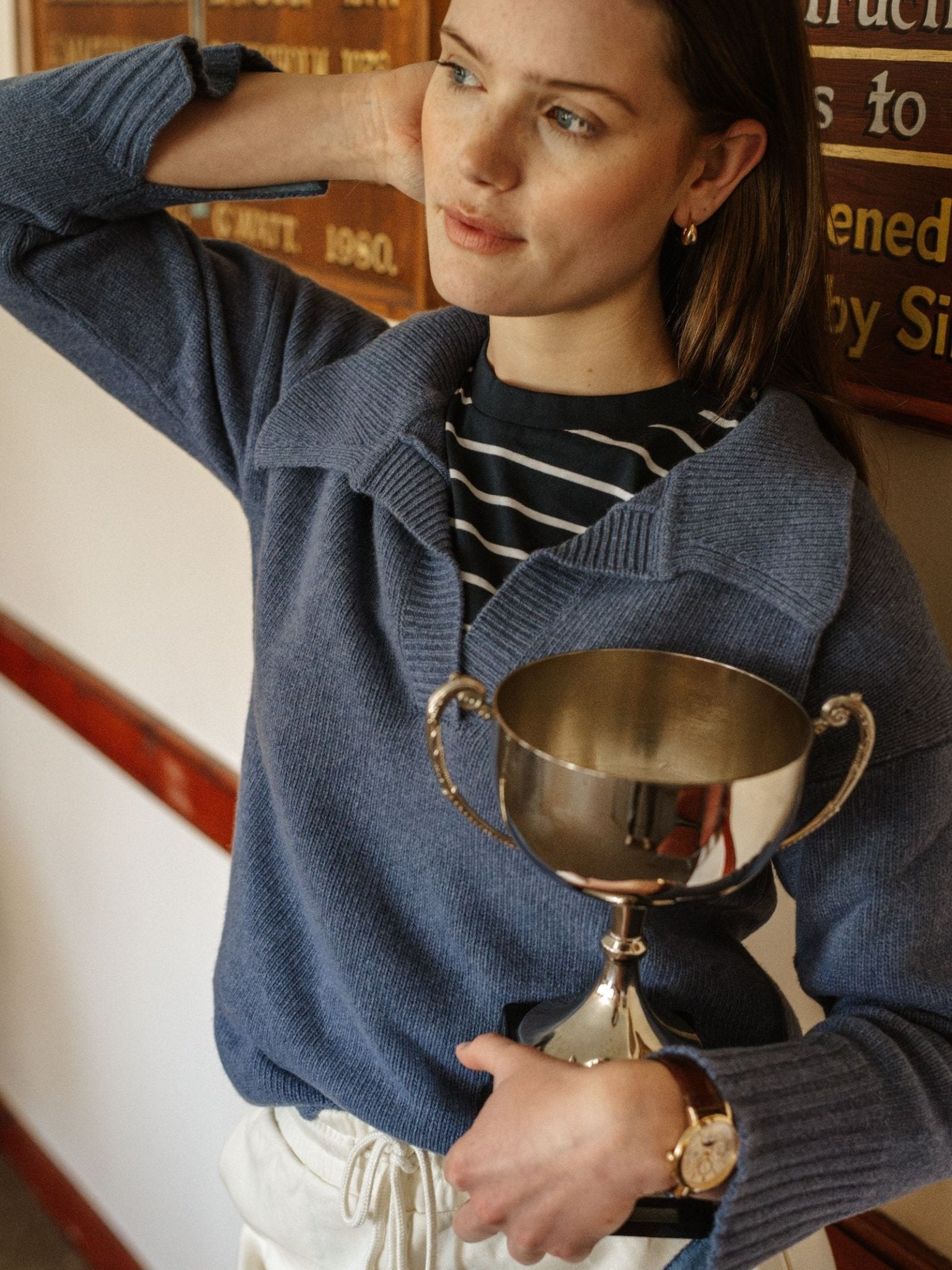 A woman with light brown hair wears a Campbell's of Beauly Merino Cashmere Shirt Neck Jumper in blue and a striped shirt, holding a silver trophy indoors near a wooden plaque. She wears a watch and gazes thoughtfully to the side.