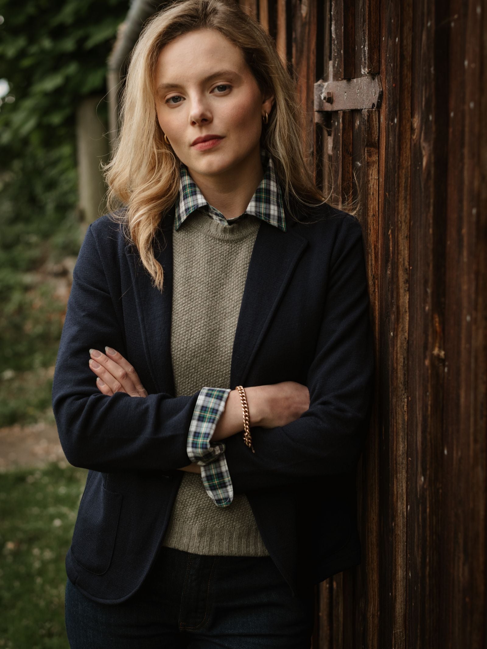 A confident blonde woman stands outside against a wooden wall, arms crossed, wearing a plaid shirt under a gray sweater and the navy Merino Blazer by Campbell's of Beauly. Green foliage is visible in the background.