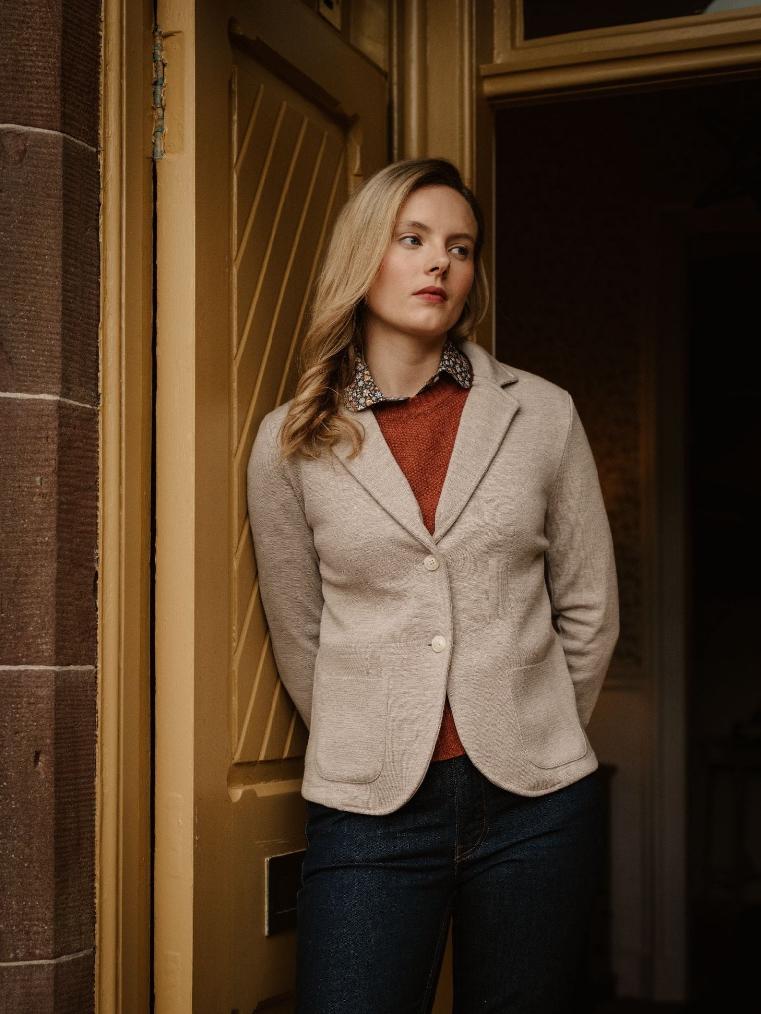A blonde woman in a beige Campbell's of Beauly Merino Blazer, rust sweater, and dark jeans stands in a doorway with her hands behind her back, gazing thoughtfully into the distance.