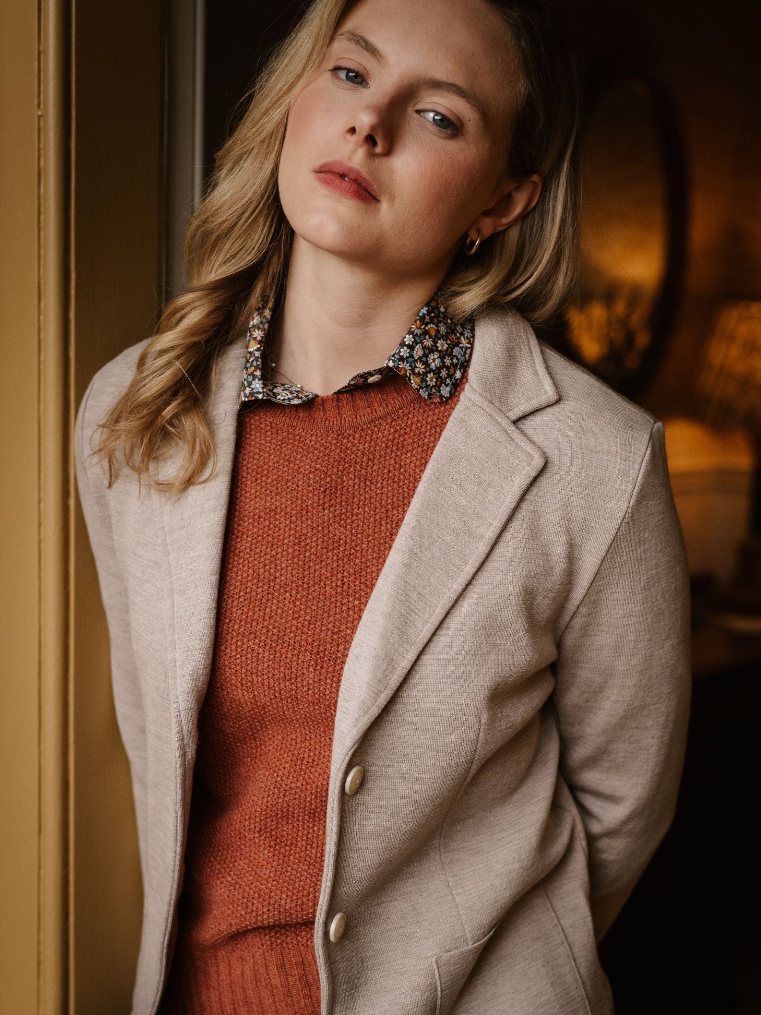 A woman with wavy blonde hair wears the Campbell's of Beauly Merino Blazer in beige over a burnt orange sweater and floral collared shirt, standing indoors and looking slightly upward with a neutral expression.