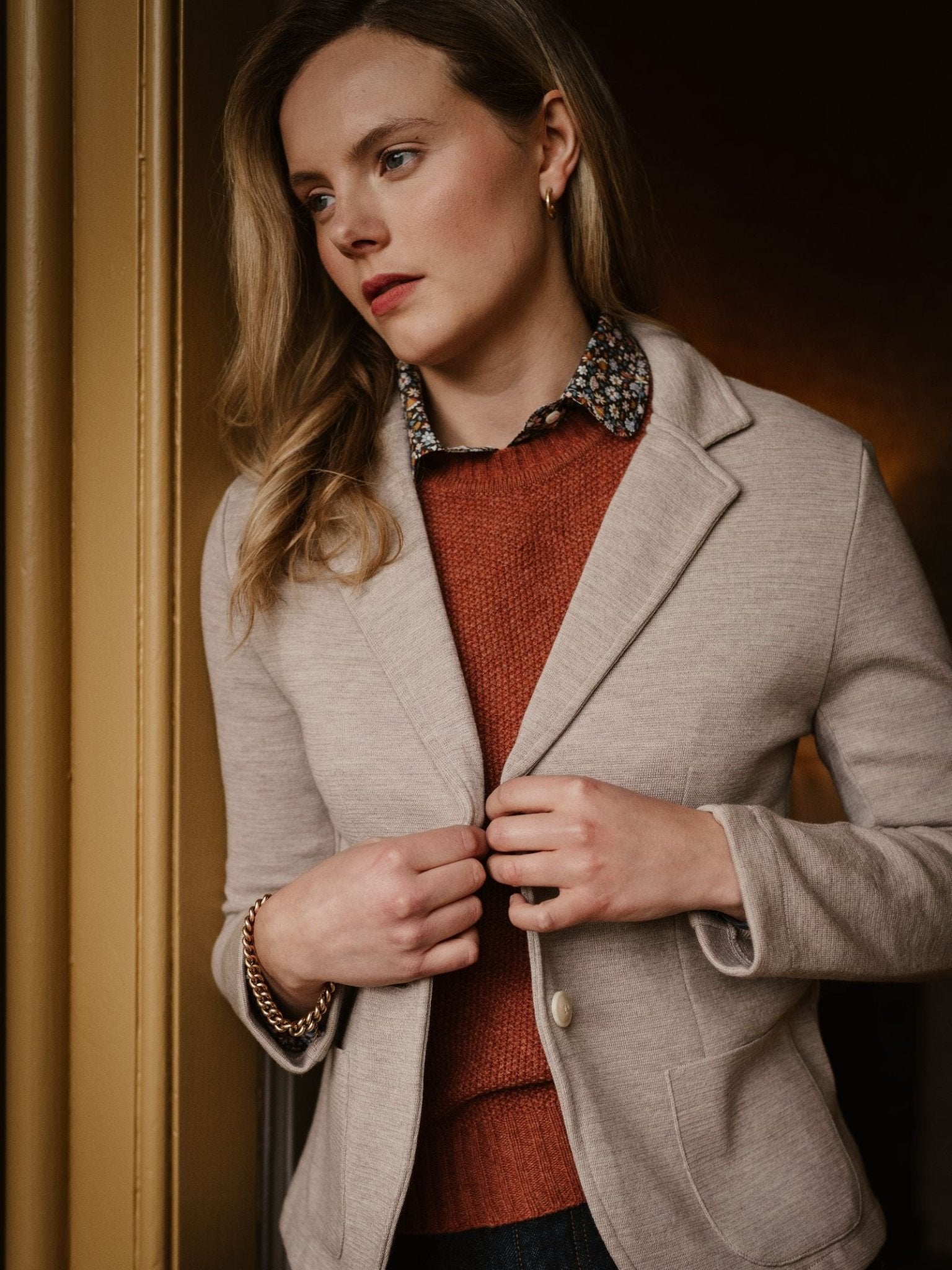A woman with long blonde hair stands indoors, thoughtfully gazing to the side while holding her Campbell's of Beauly Merino Blazer, worn over a rust sweater and floral-collared shirt.