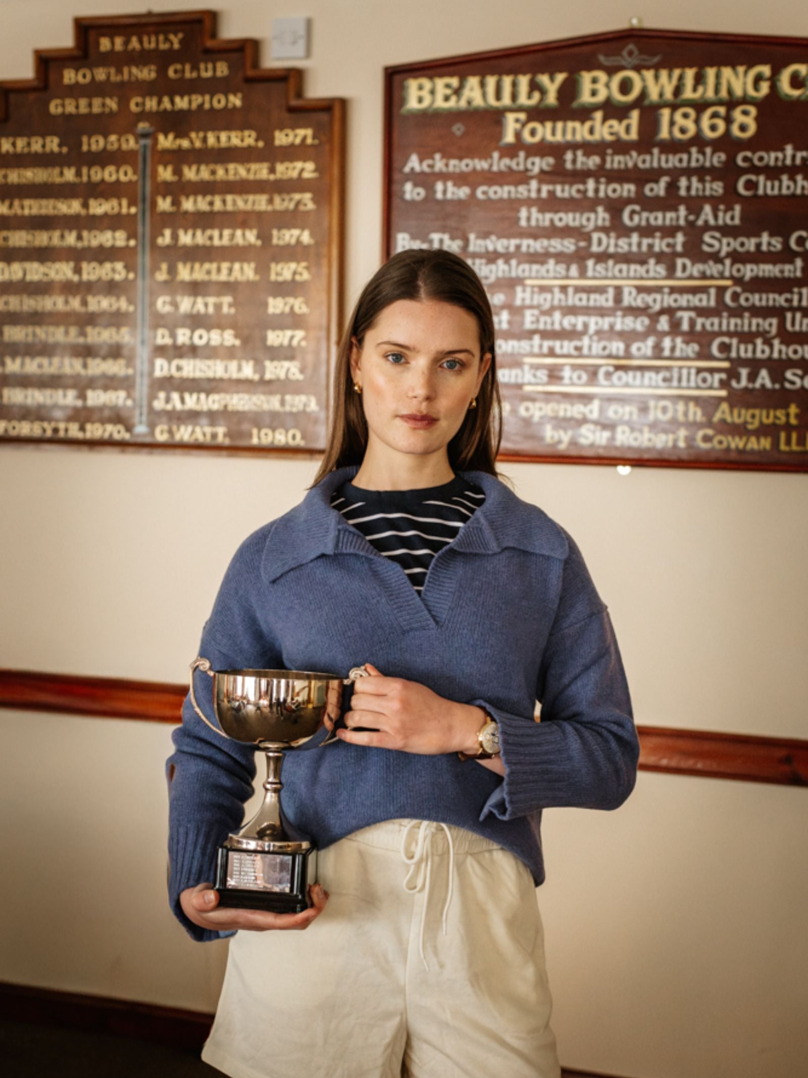A young woman with straight brown hair stands indoors, holding a silver trophy and wearing a Campbell's of Beauly Merino Cashmere Shirt Neck Jumper over a striped shirt and cream shorts, looking confidently at the camera.