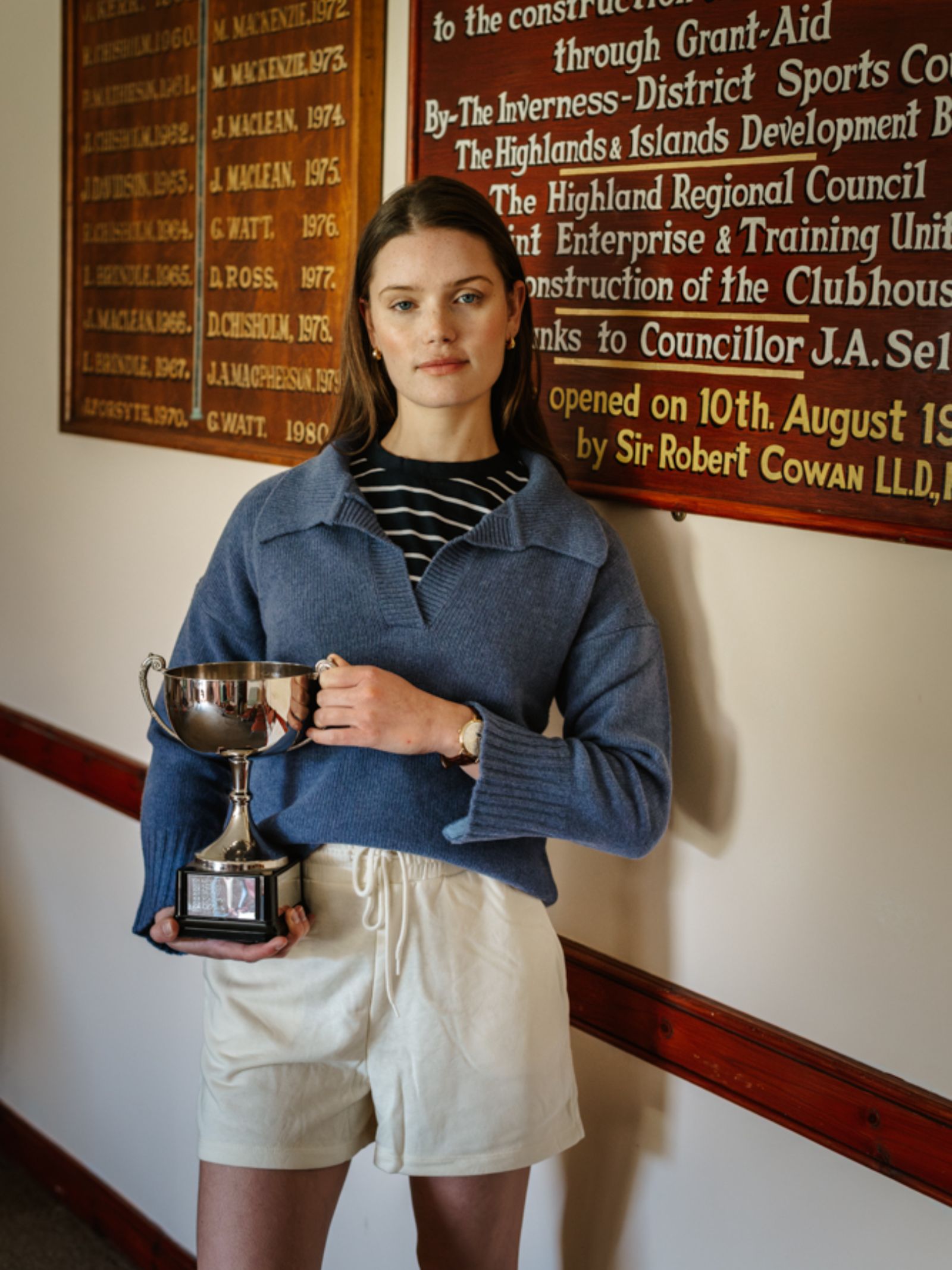 A young woman with straight brown hair wears a Campbell's of Beauly Merino Cashmere Shirt Neck Jumper over a striped shirt and white shorts, holding a silver trophy indoors by a wooden plaque and looking confidently at the camera.