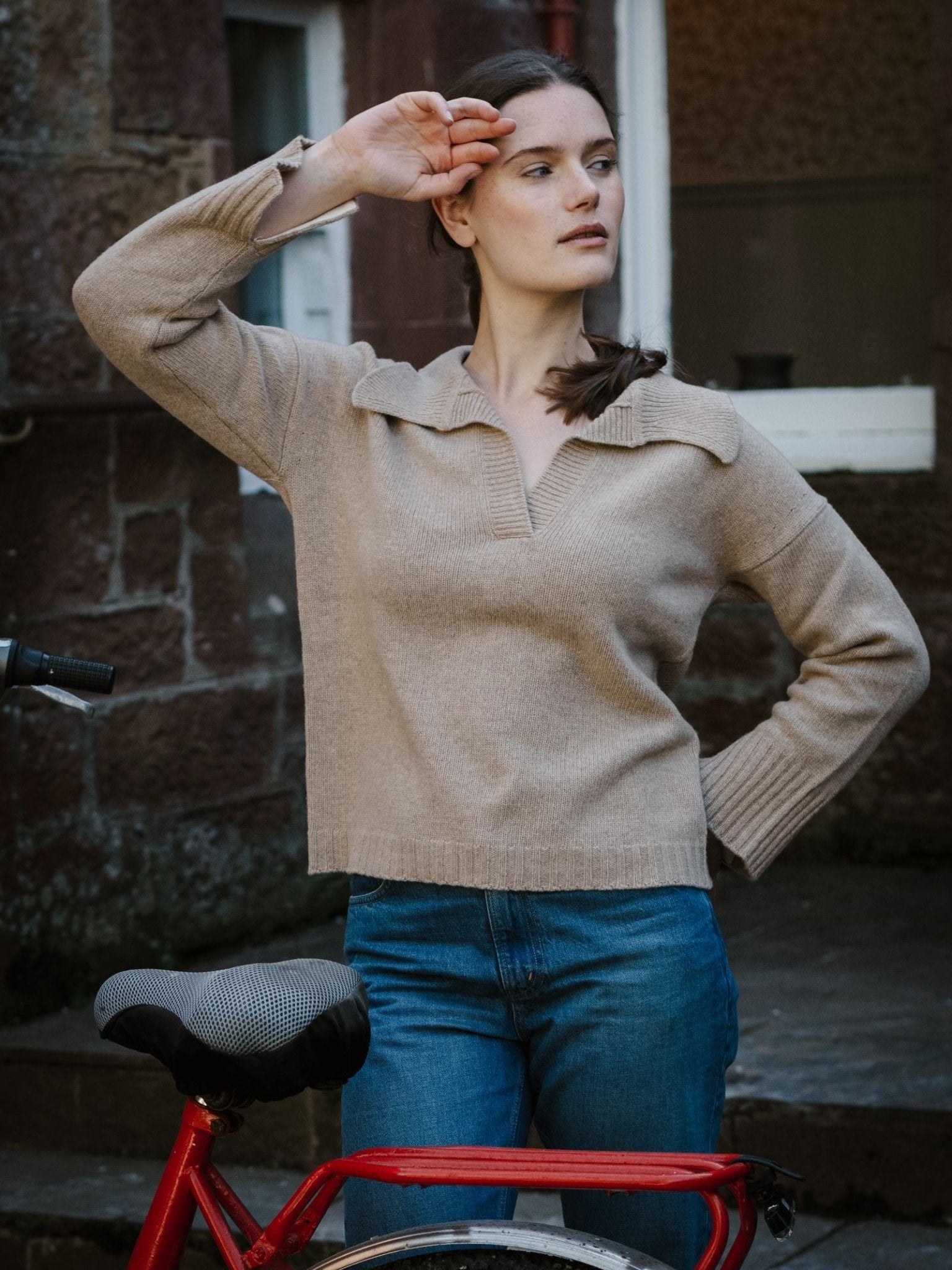 A young woman in a Campbell's of Beauly Merino Cashmere Shirt Neck Jumper and blue jeans stands by a red bike, raising her right hand to her forehead, with a brick wall and window behind her.