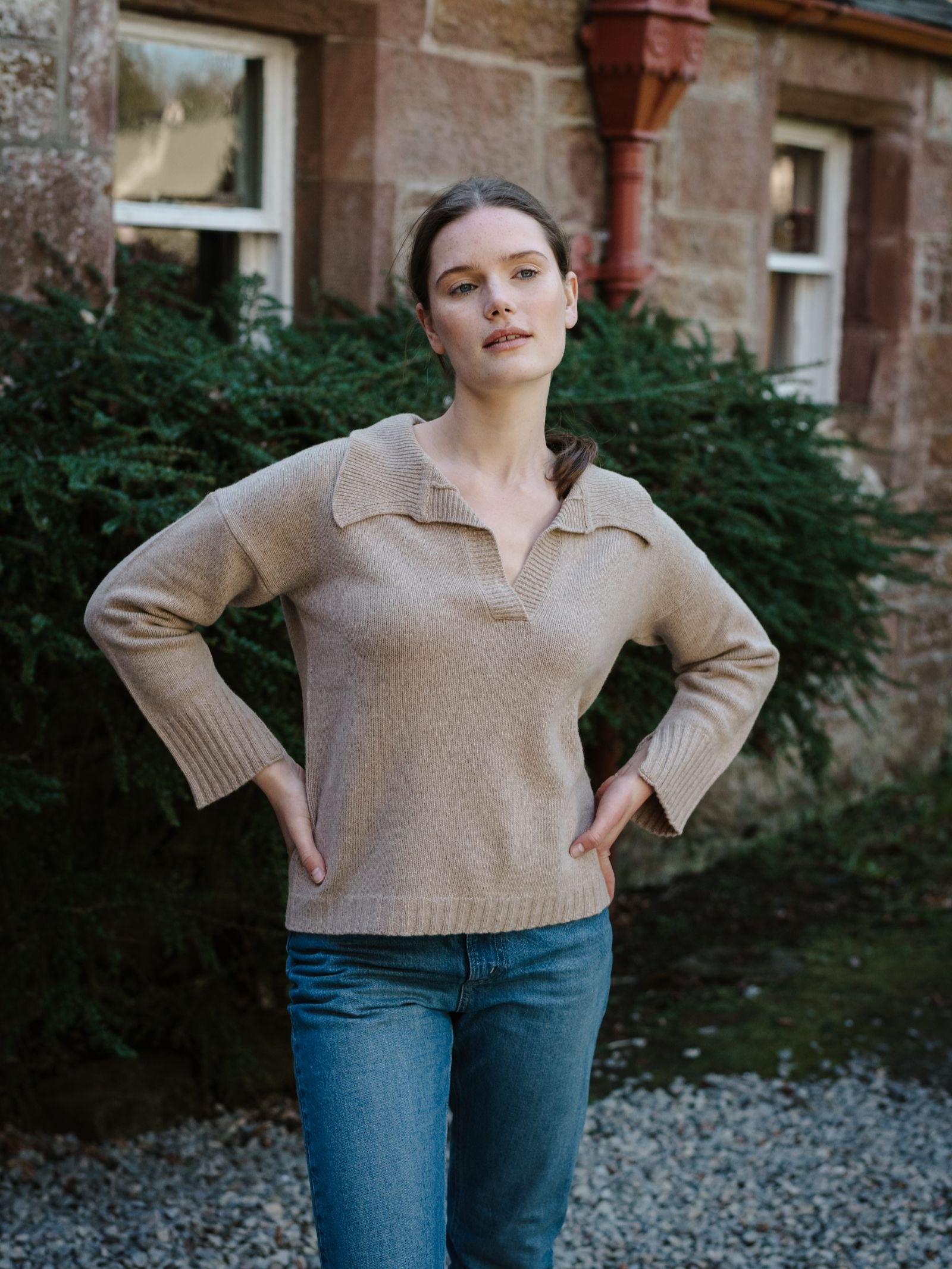 A woman with light skin and brown hair stands outdoors in front of a stone building and greenery, hands on hips, wearing a Campbell's of Beauly Merino Cashmere Shirt Neck Jumper and blue jeans, looking slightly upward.