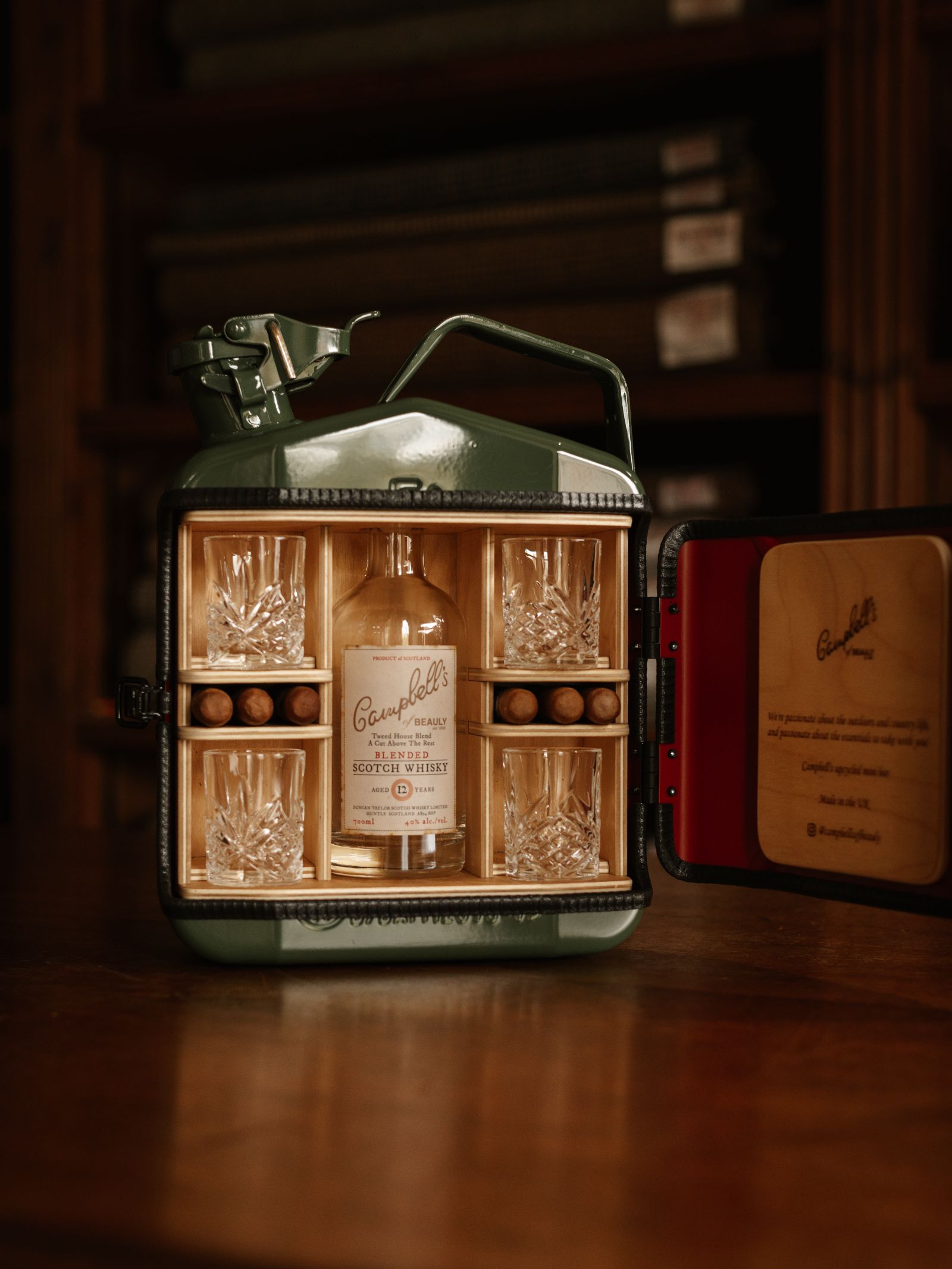 The Mini Connoisseur Upcycled Mini Bar from Campbell's of Beauly, a repurposed jerry can gift box, contains a whiskey bottle, four glasses, and six stones—shown on a wooden table with bookshelves in the background.