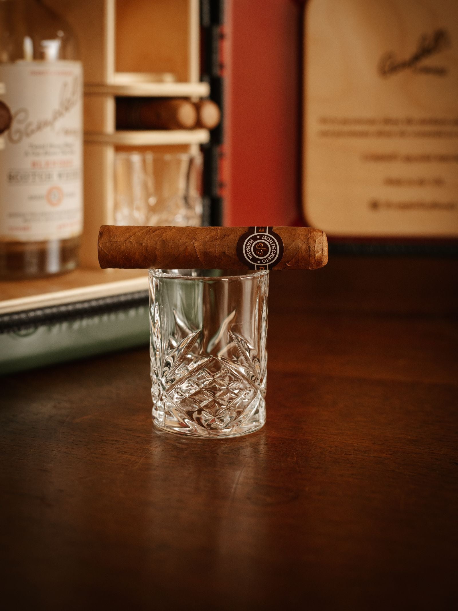 A cigar rests atop an empty crystal glass on a wooden table. In the background, a bottle of Glenfiddich whisky and Campbell's of Beauly Mini Connoisseur Upcycled Mini Bar add warmth and charm to the inviting scene.
