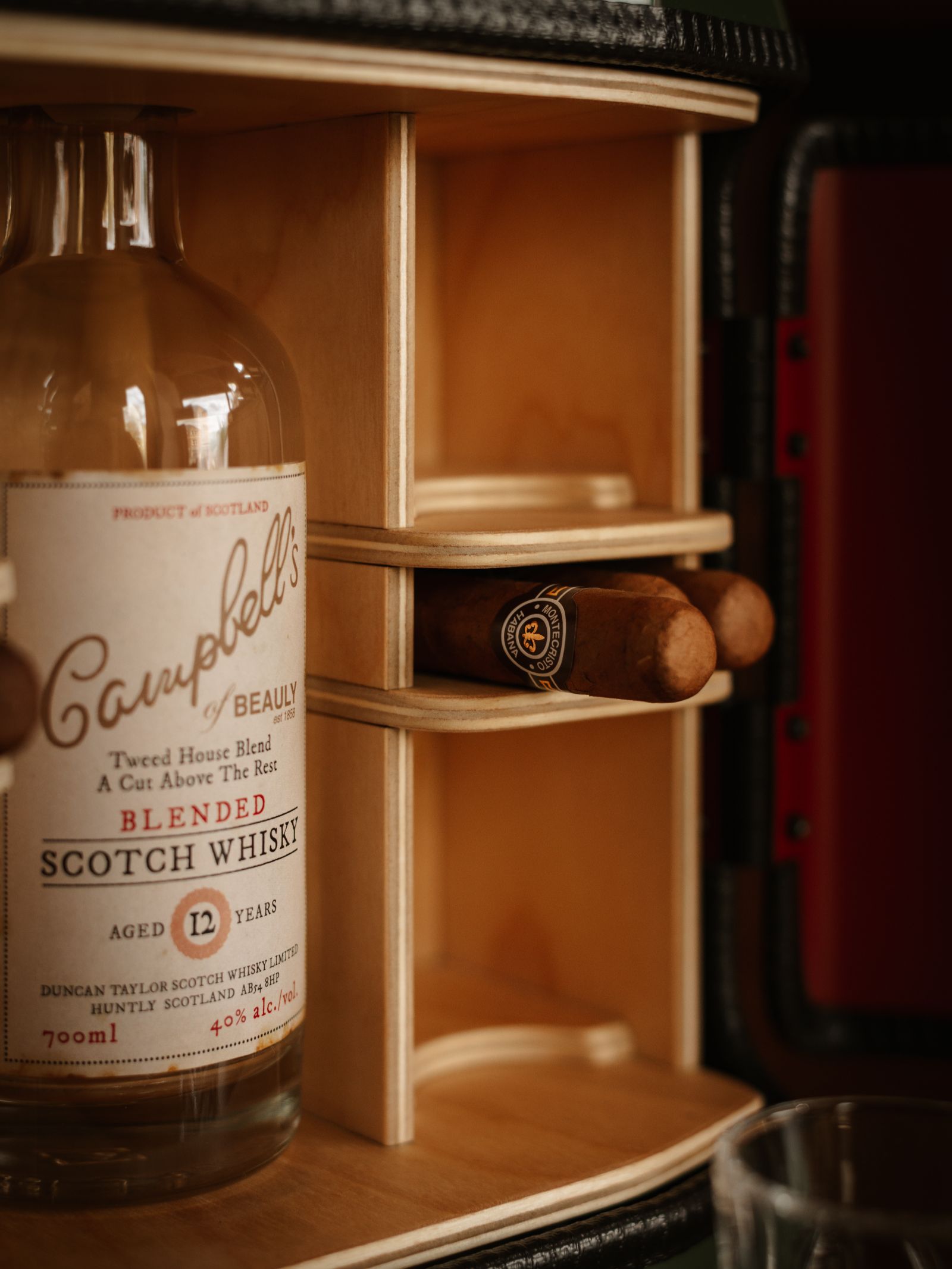 A close-up of a Mini Connoisseur Upcycled Mini Bar by Campbell’s of Beauly holds a bottle of their 12-year-old blended Scotch whisky and two cigars, with part of a glass visible in the lower right corner.