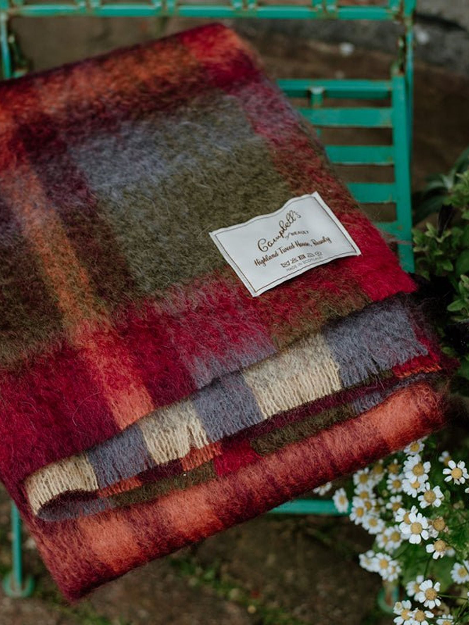 The Painterly Mohair Throw by Campbell's of Beauly, crafted in Yorkshire, rests folded on a green metal chair beside white and yellow daisies. Its soft plaid features red, purple, green, and beige tones.