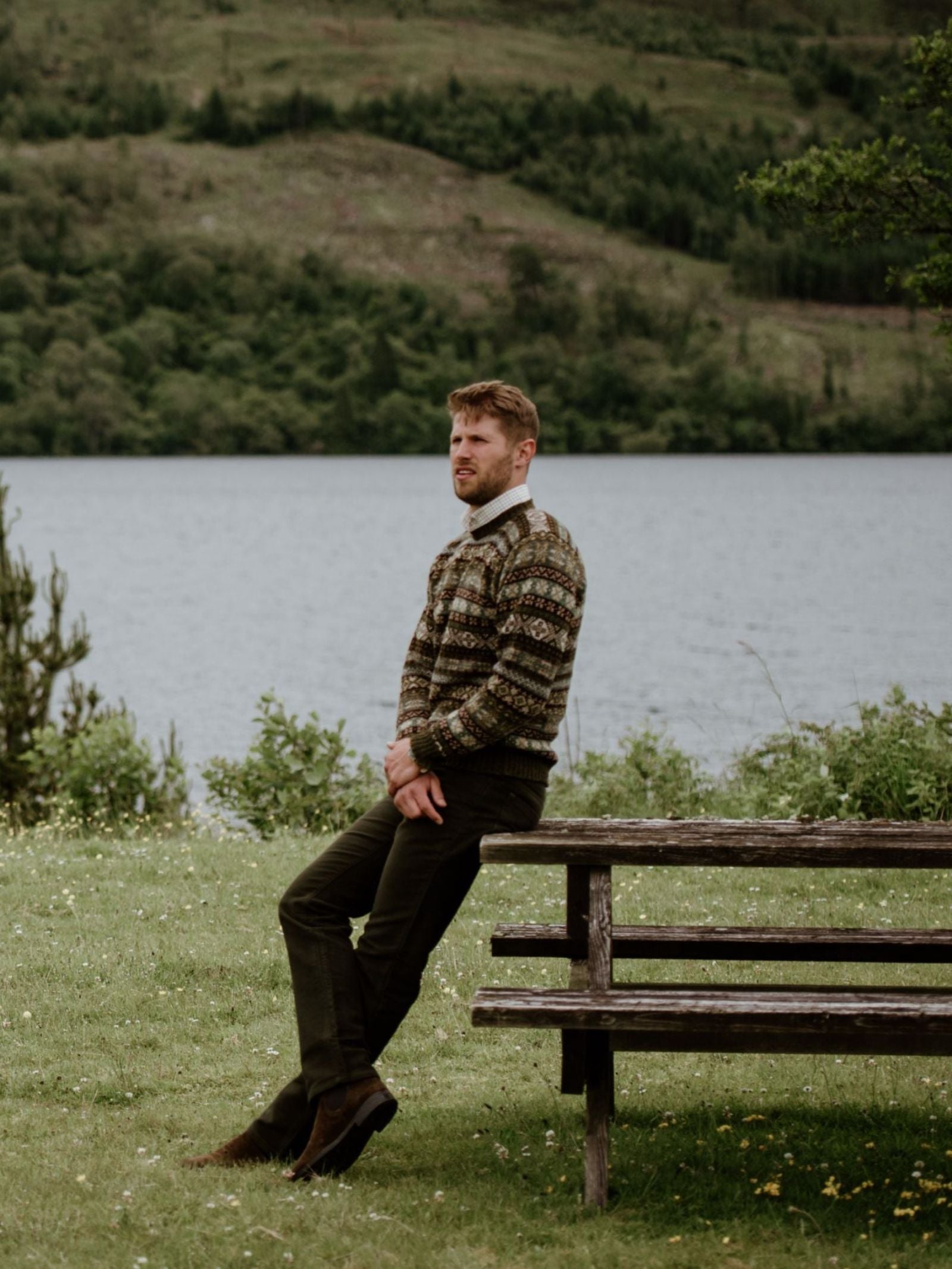 A man in a patterned sweater and Campbell's of Beauly Moleskin Jeans sits on a wooden picnic table by a lake, with green hills and trees in the background on an overcast day.