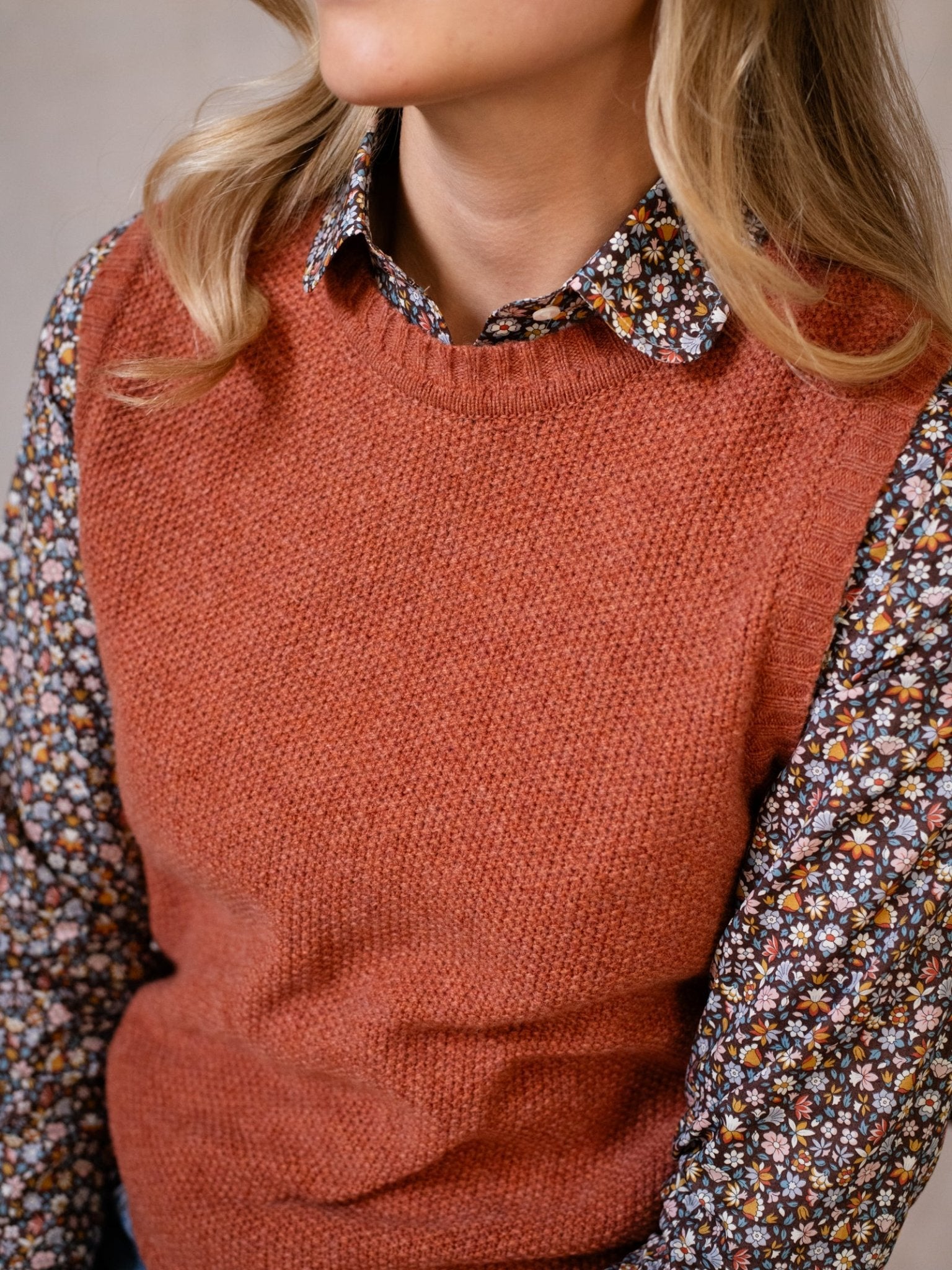 A woman with blonde hair wears the Campbell's of Beauly Moss Stitch Crew Tank, crafted in Scotland, over a floral button-up shirt. Her face is partially out of frame as she sits with her hands in her lap.