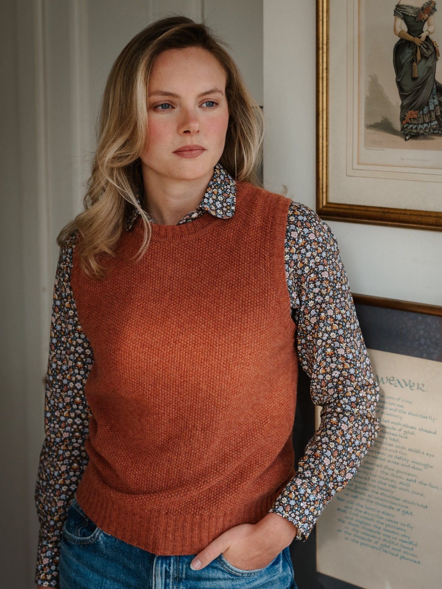 A woman with blonde hair wears the Campbell's of Beauly Moss Stitch Crew Tank over a floral shirt, paired with jeans, standing indoors beside framed artwork.