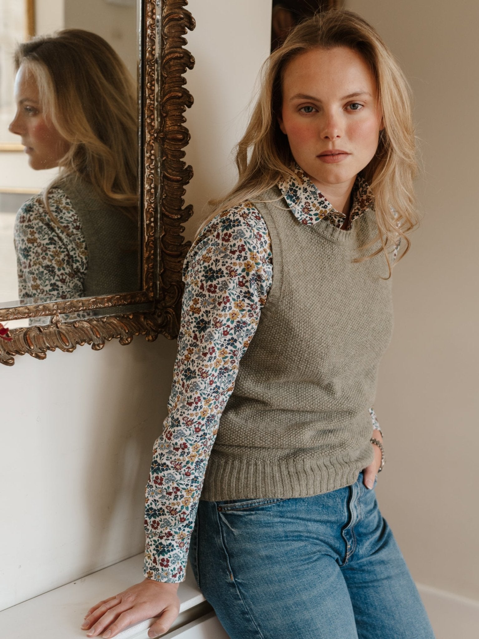 A woman with long blonde hair, in a floral shirt and Campbell's of Beauly Moss Stitch Crew Tank, leans against a white wall by an ornate mirror in blue jeans, looking calmly at the camera.